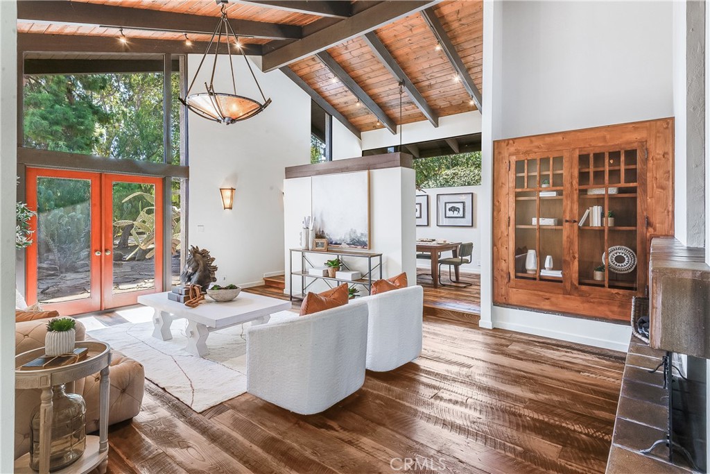 Living room with vaulted wood ceiling, exposed beams, polished floors, and red-orange doors opening toward trees.