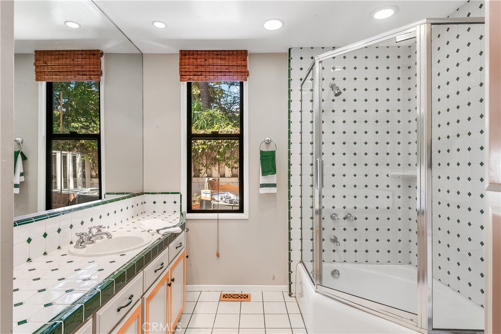 Bathroom with a tiled vanity, green-accented tile, two windows, and a glass-enclosed tub and shower.
