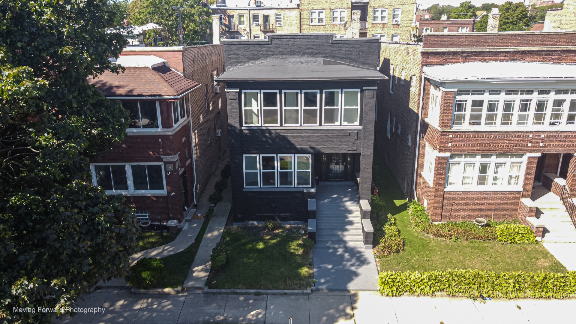 Dark gray brick home with large front windows, broad stairs, and a small lawn.