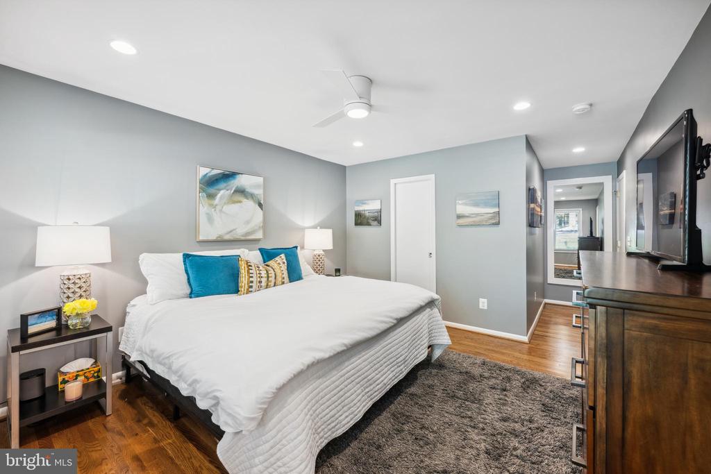 Staged bedroom with a white bed, teal pillows, hardwood floors, and a wall-mounted television.