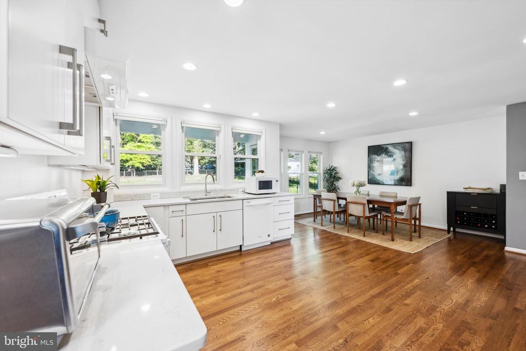 Open white kitchen and dining area with hardwood floors, large windows, and a table on a rug.