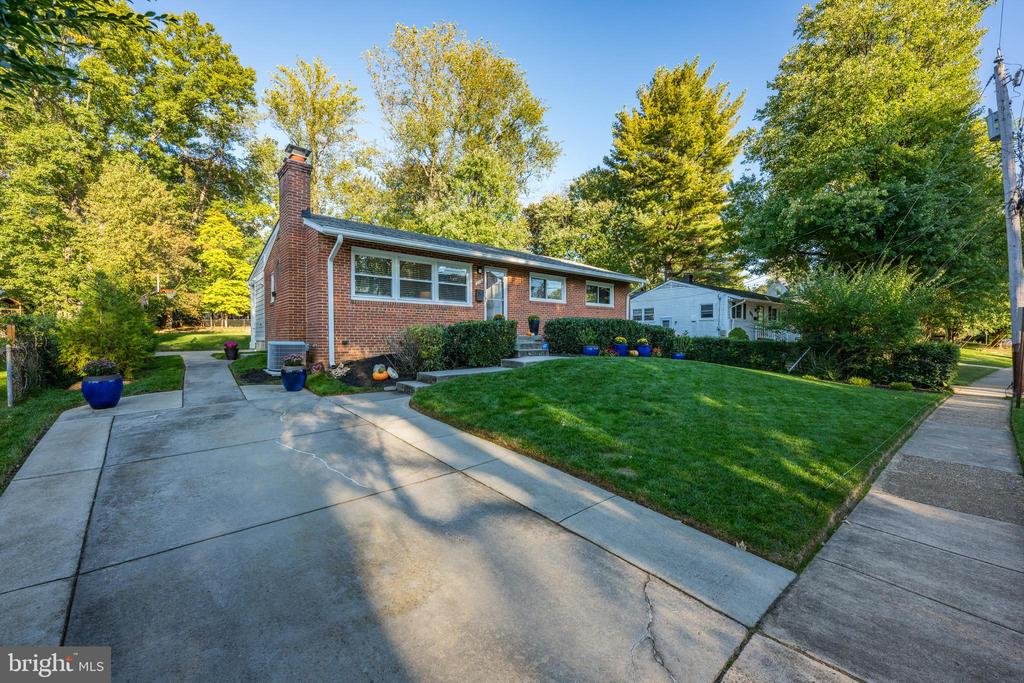 Red brick ranch home with a broad concrete driveway, green lawn, and mature trees.