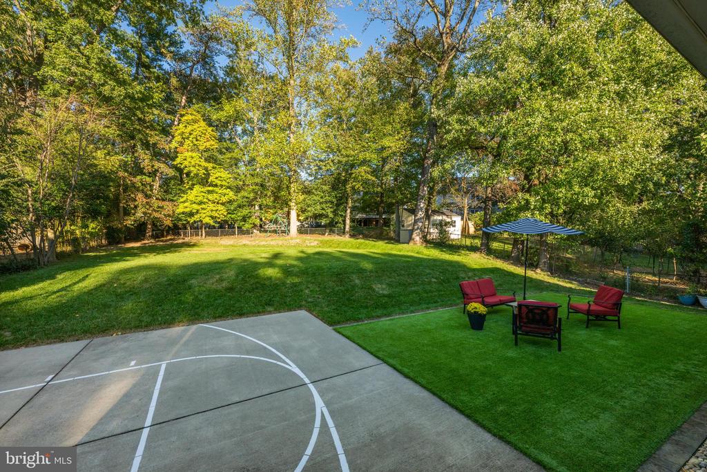 Wooded backyard with a basketball court and a red-chair seating area beneath a blue umbrella.