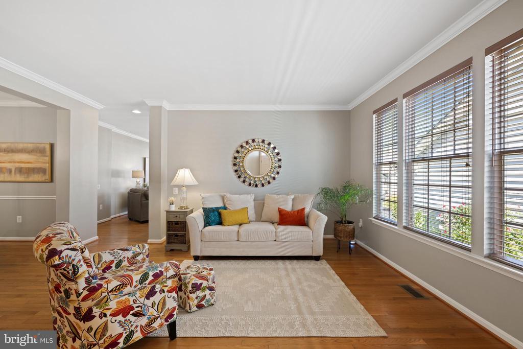 Living room with a cream sofa, patterned armchair, hardwood floors, and tall windows with blinds.