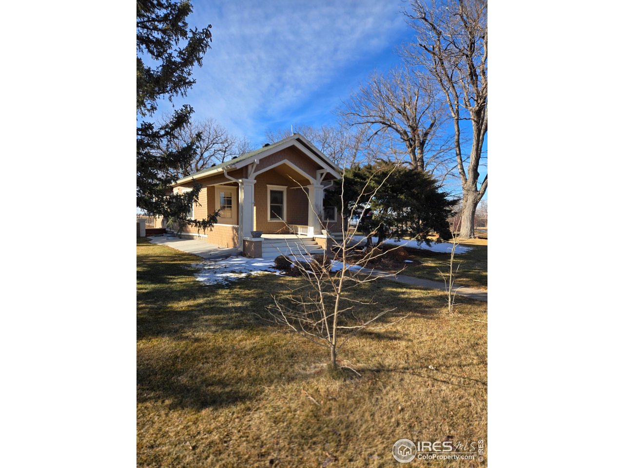 Tan house with a covered front porch, mature trees, and patches of snow in the yard.