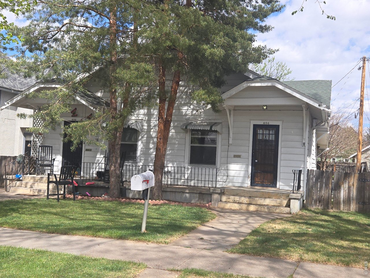 Two adjoining light-colored homes with covered porches and mature pine trees in front.