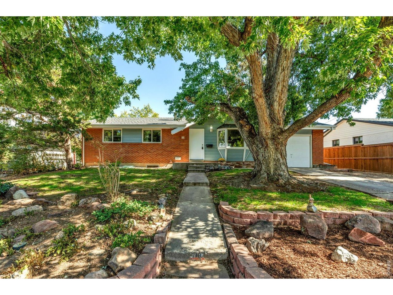 Single-story brick house with an attached garage, shaded by a large tree over a landscaped front yard.