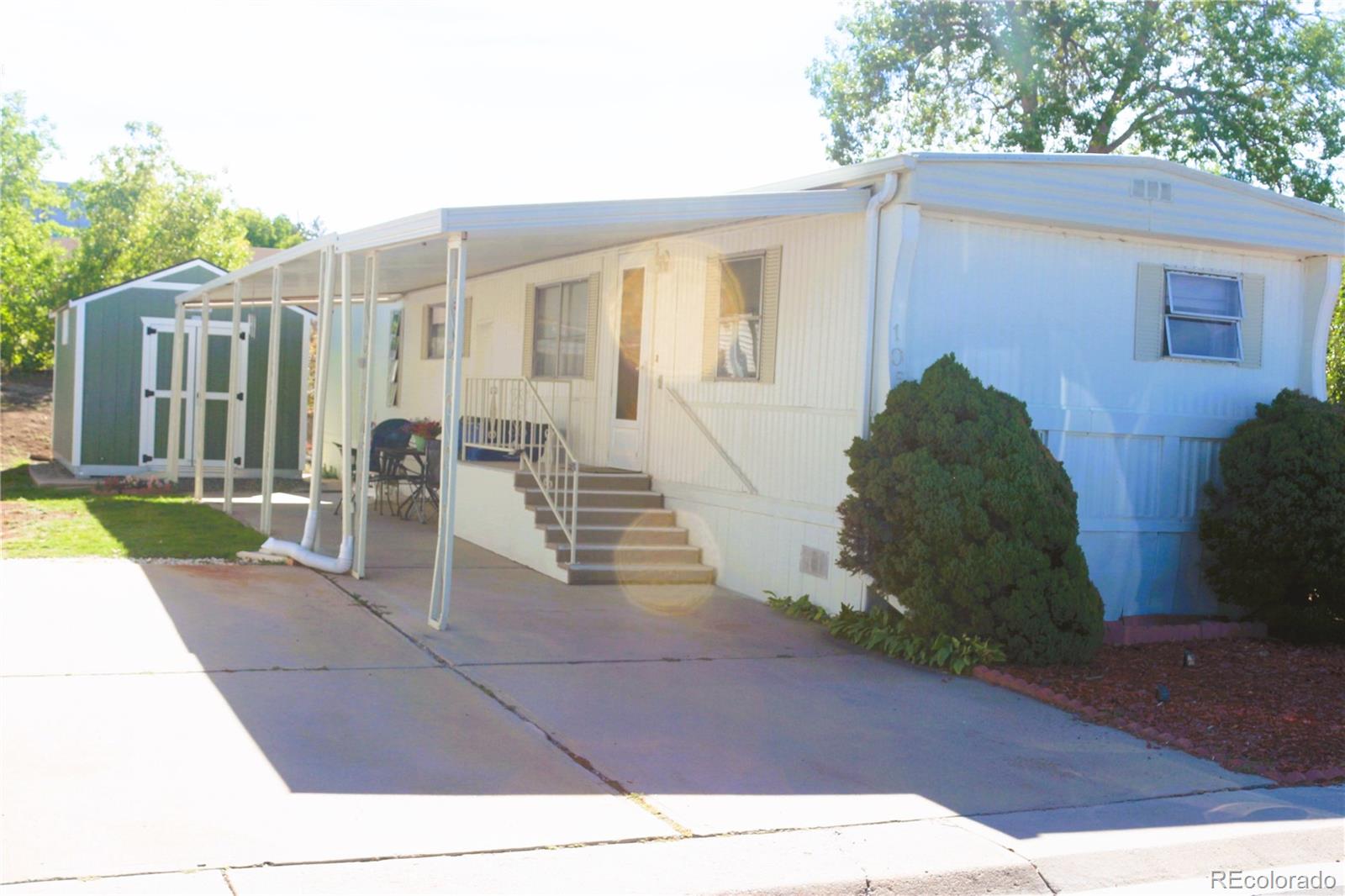 White manufactured home with a covered porch, front steps, and a green storage shed beside the driveway.