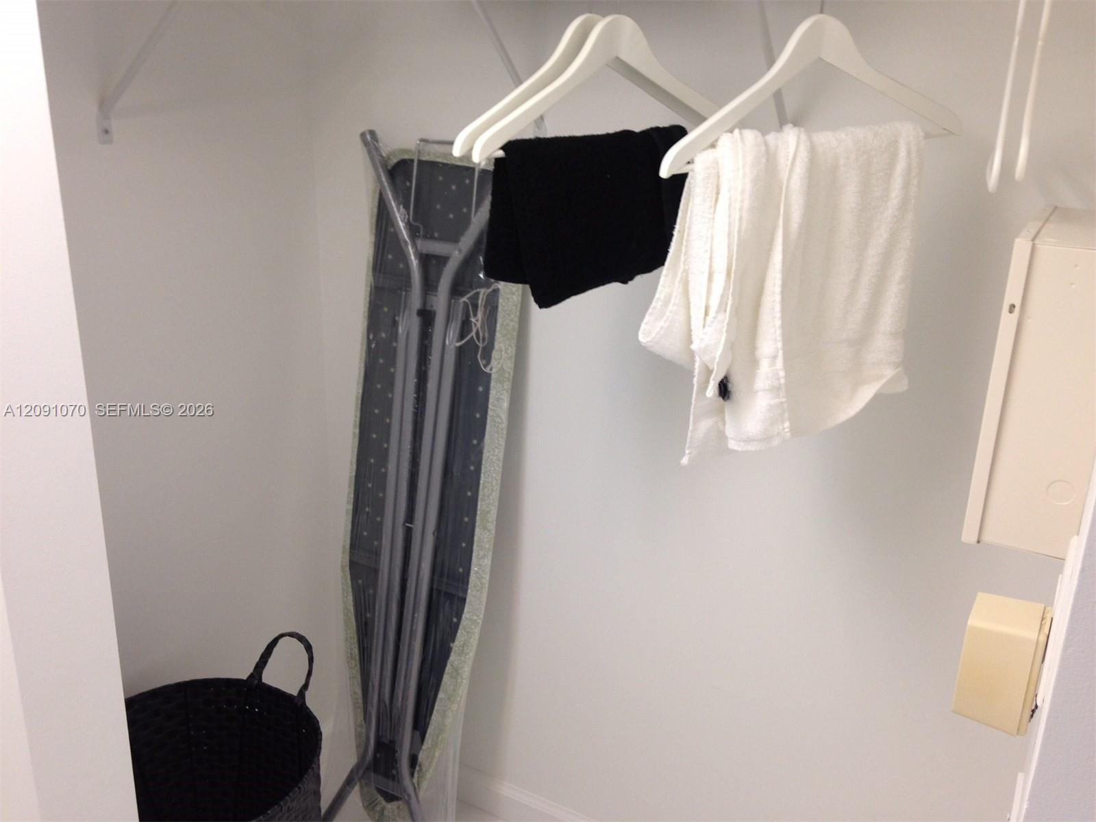 Closet with hanging towels, an ironing board, and a black laundry basket.