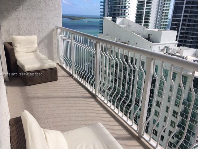 Furnished high-rise balcony with white lounge chairs overlooking the bay and neighboring towers.