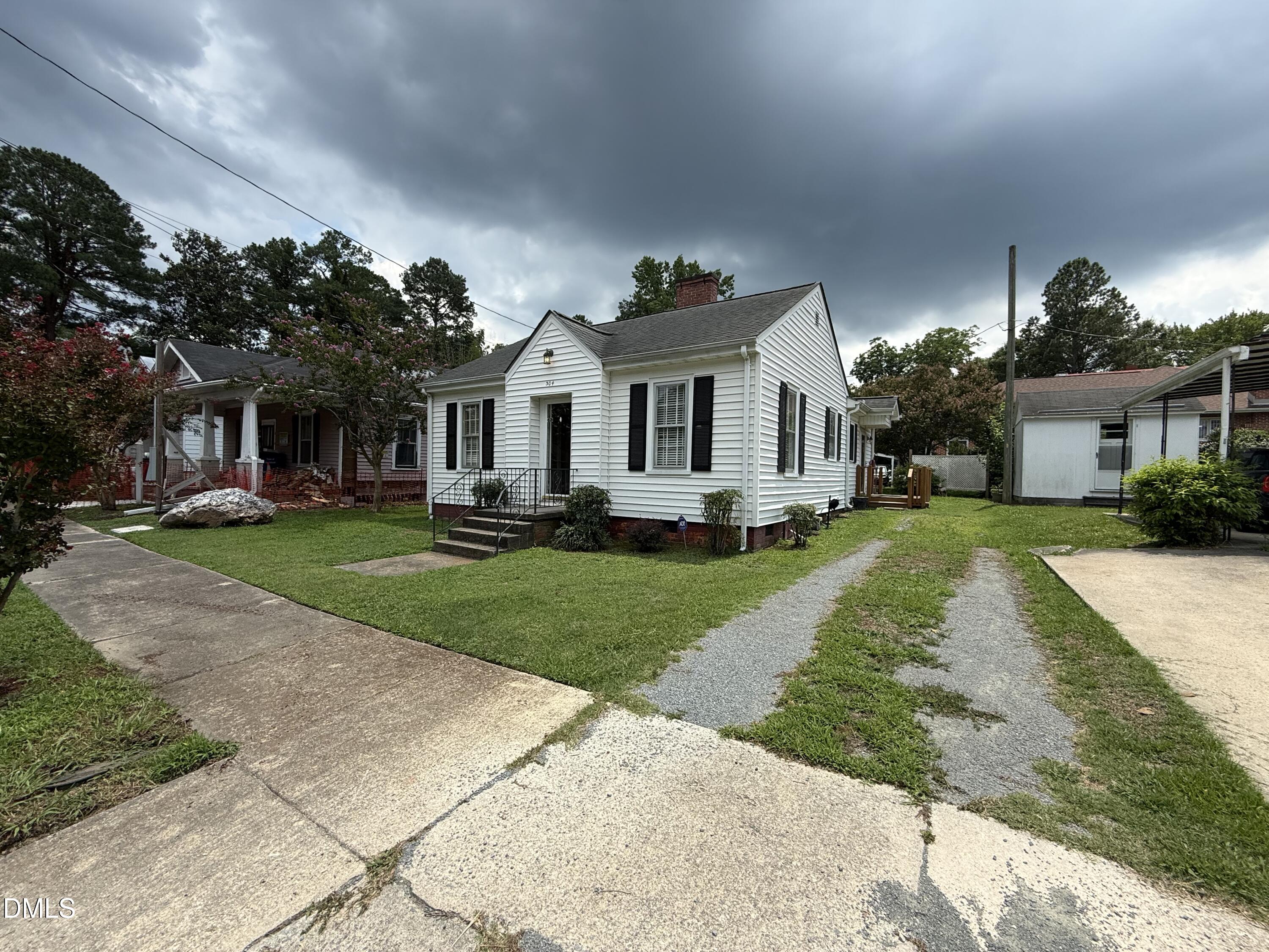 White-sided home with black shutters, front steps, lawn, and gravel driveway beneath storm clouds.
