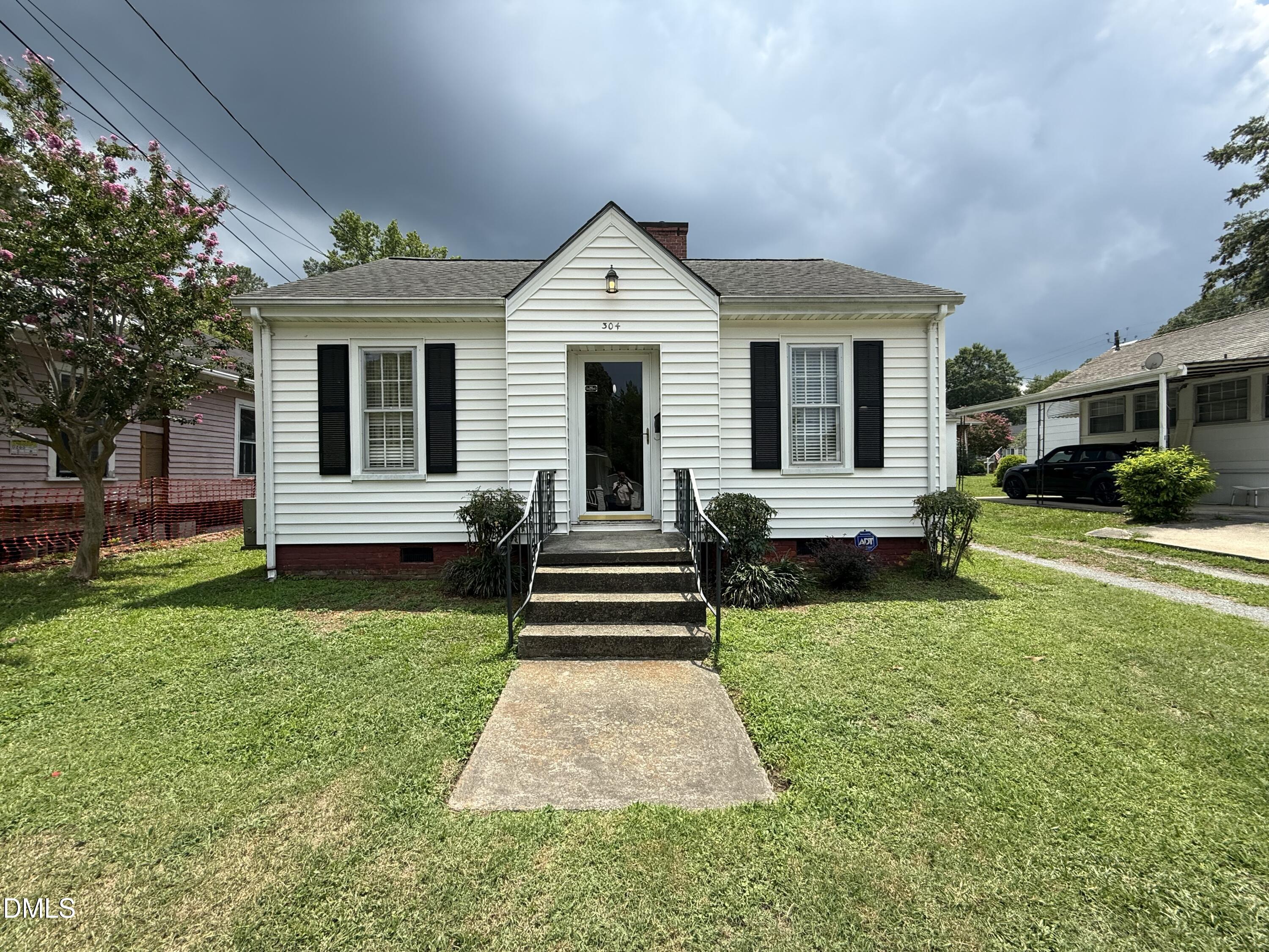 White-sided house with black shutters, a central gabled entry, concrete steps, and a green front lawn.