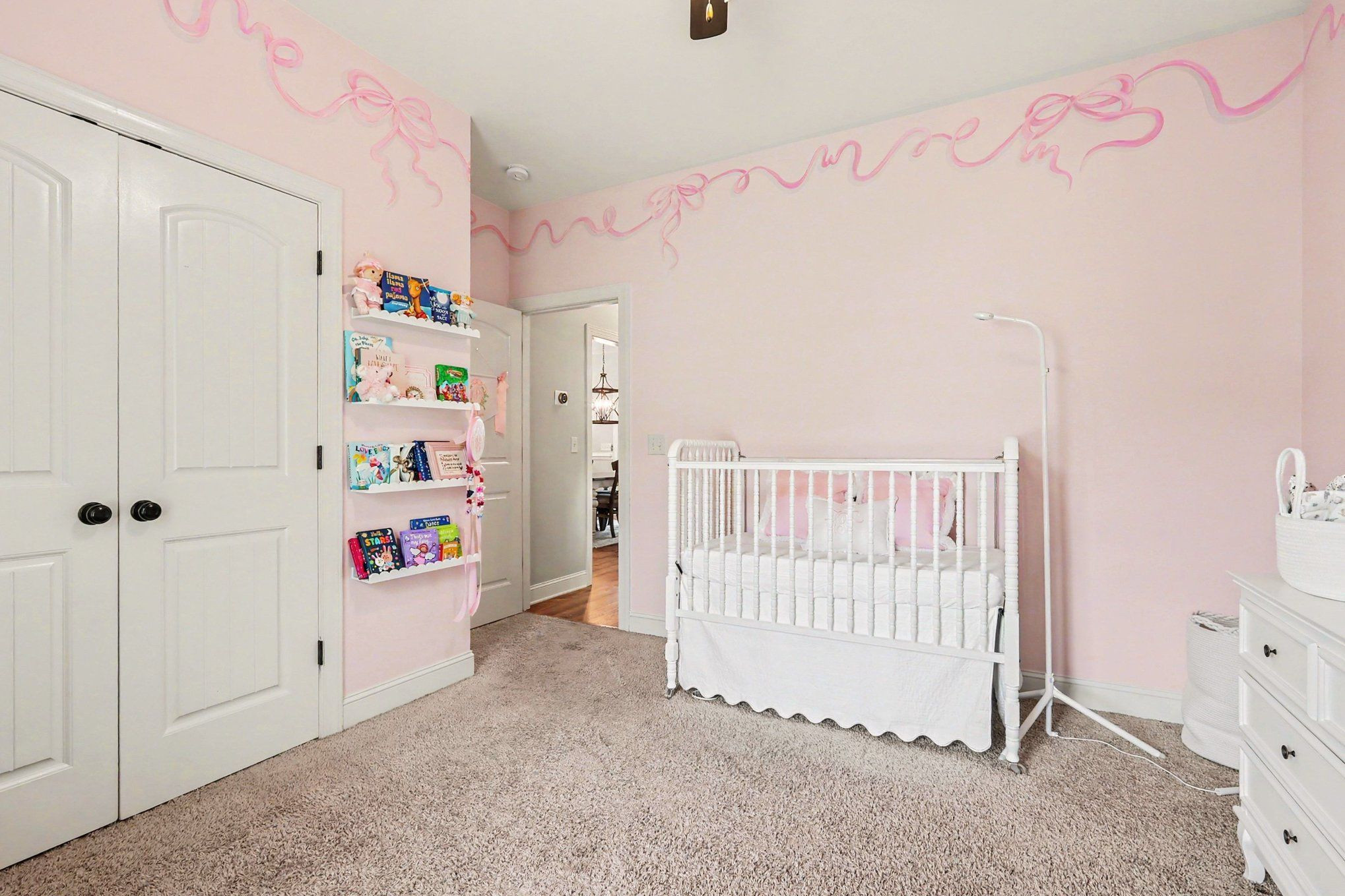 Pink nursery with a white crib, book shelves, and ribbon designs on the walls.