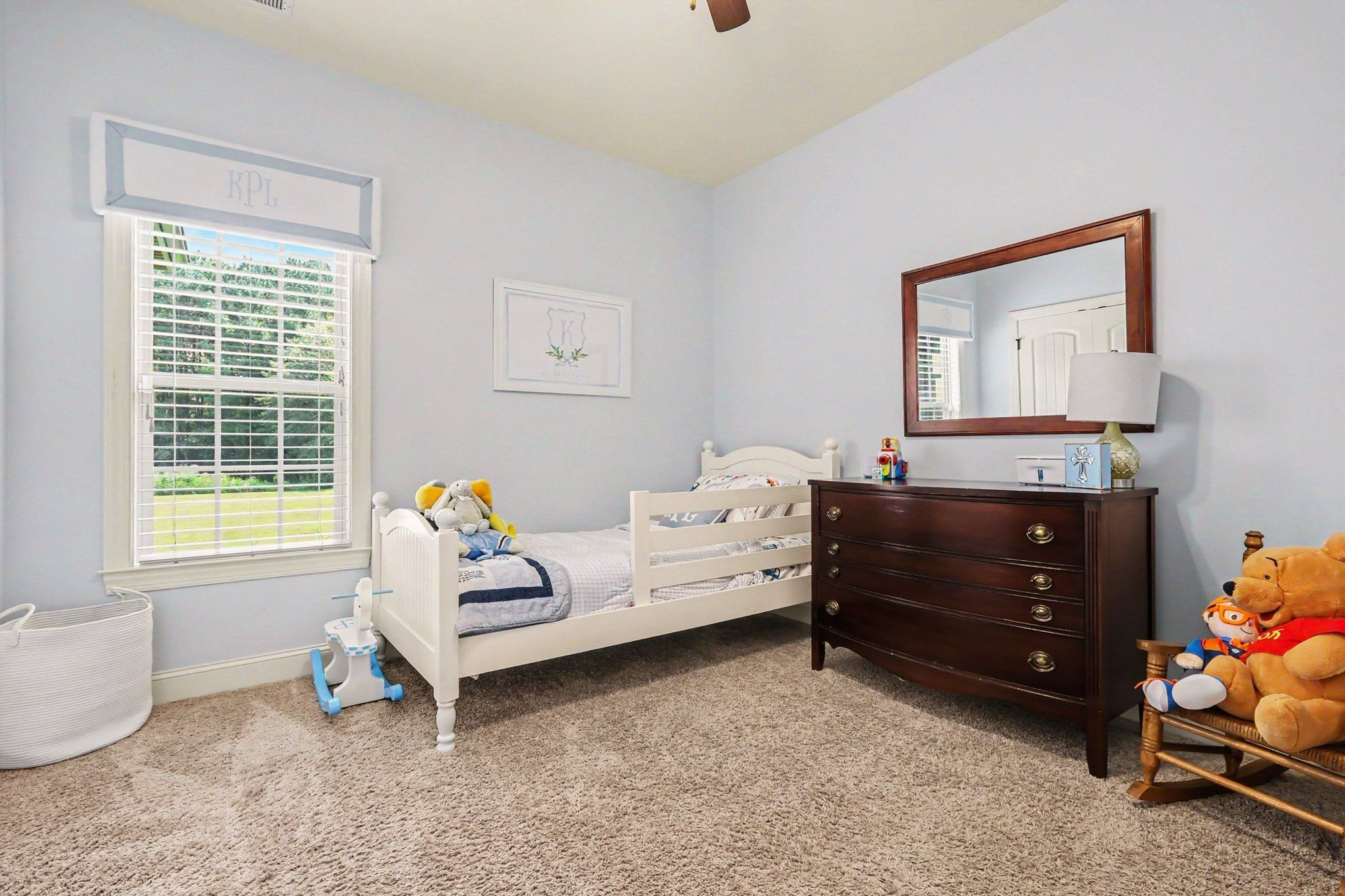 Child's bedroom with a white bed, plush toys, dark wood dresser, and a large window overlooking a lawn.