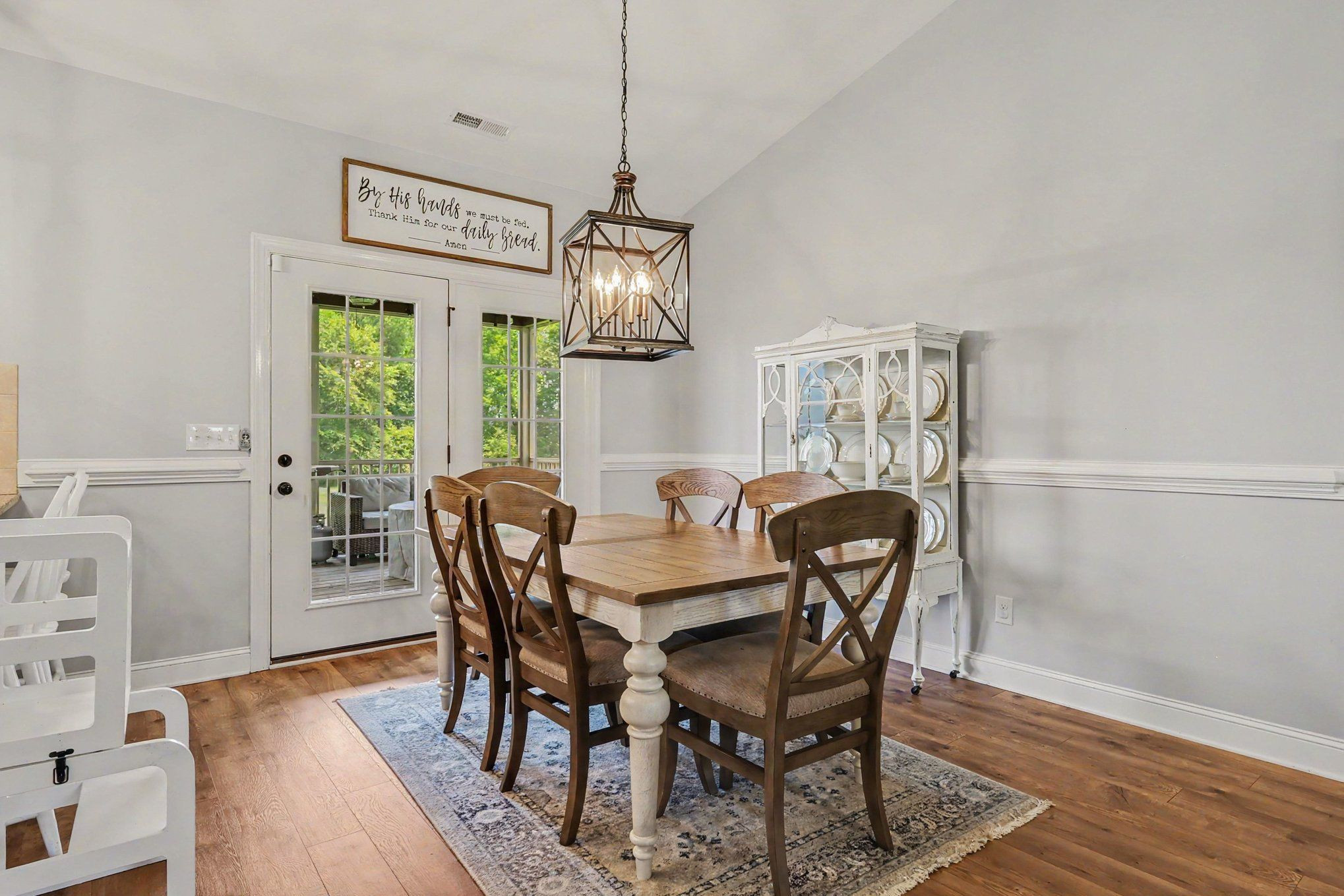 Dining room with a wooden table, cross-back chairs, china cabinet, and glass door overlooking greenery.