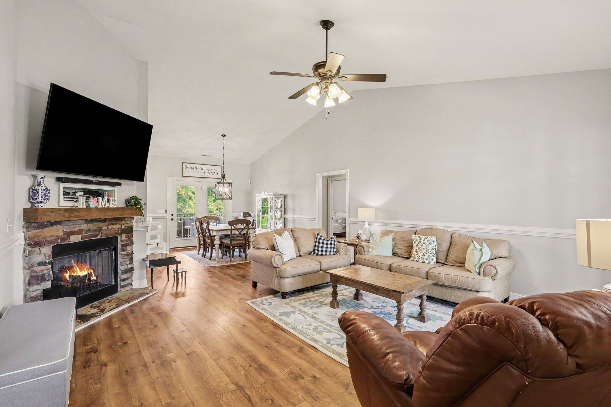 Living room with a lit stone fireplace, vaulted ceiling, wood floors, and an adjoining dining area.