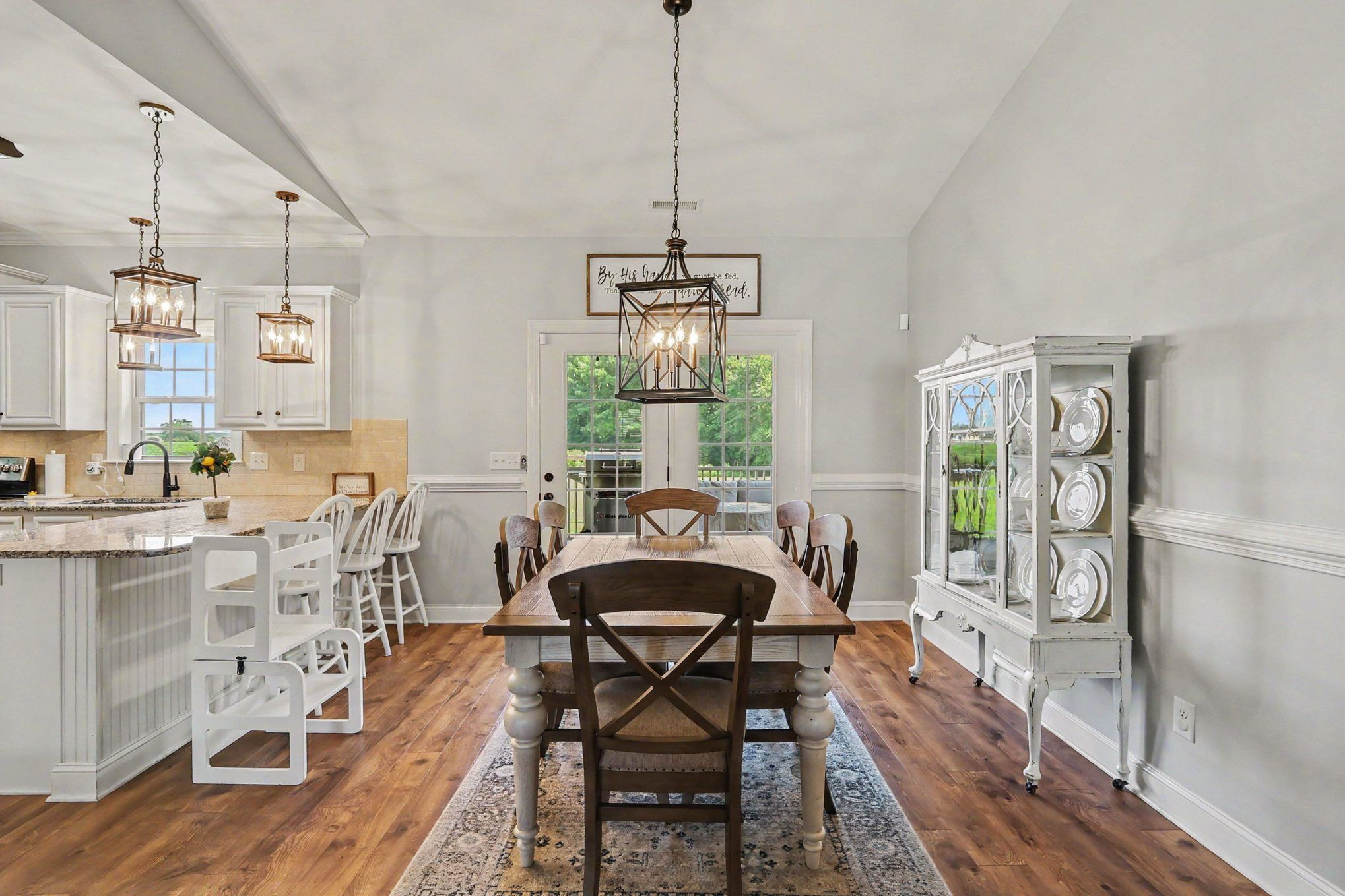 Dining room with a long wooden table, pendant lights, and glass doors beside a white kitchen.