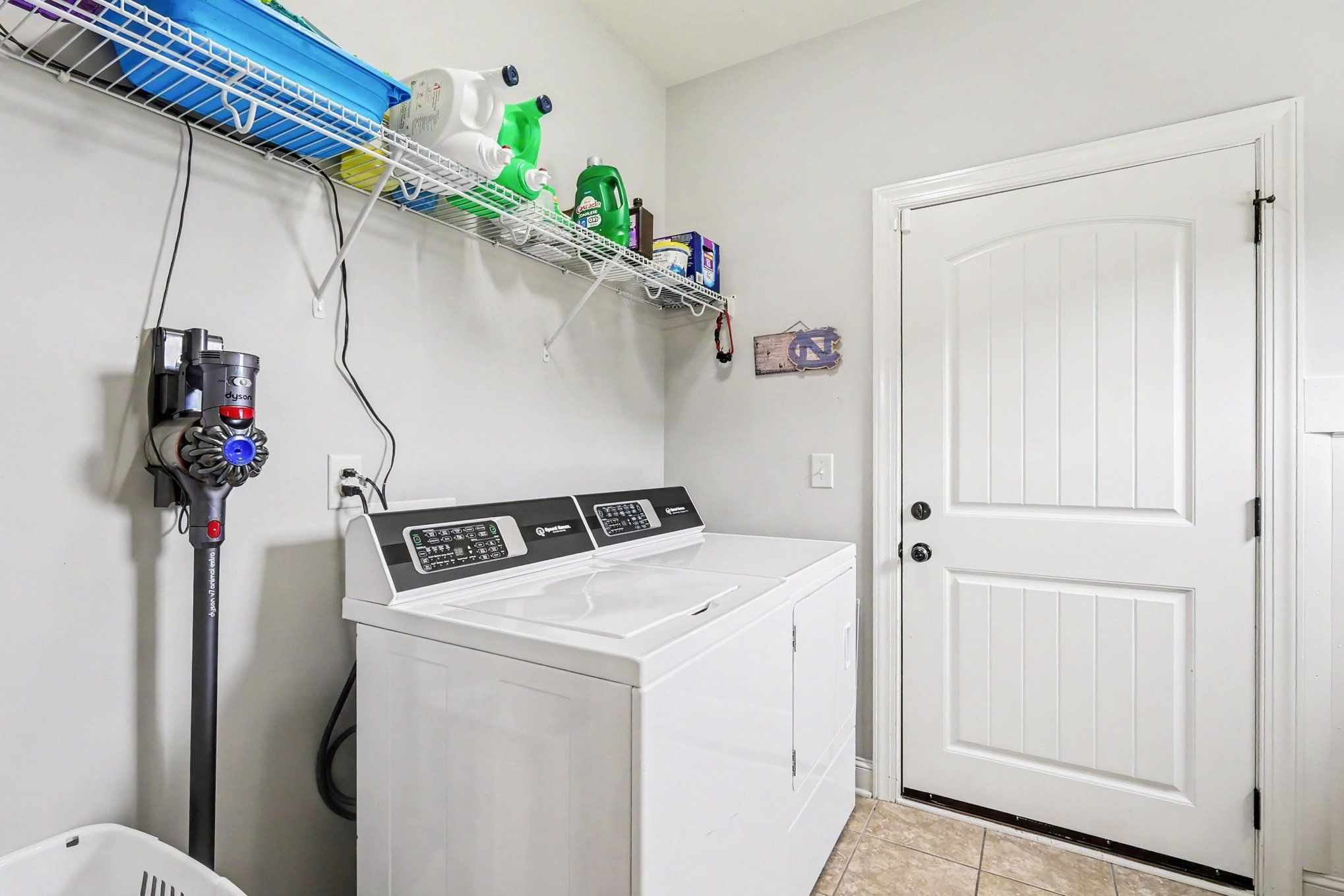 Laundry room with white top-loading washer and dryer, wire storage shelf, wall-mounted vacuum, and paneled door.