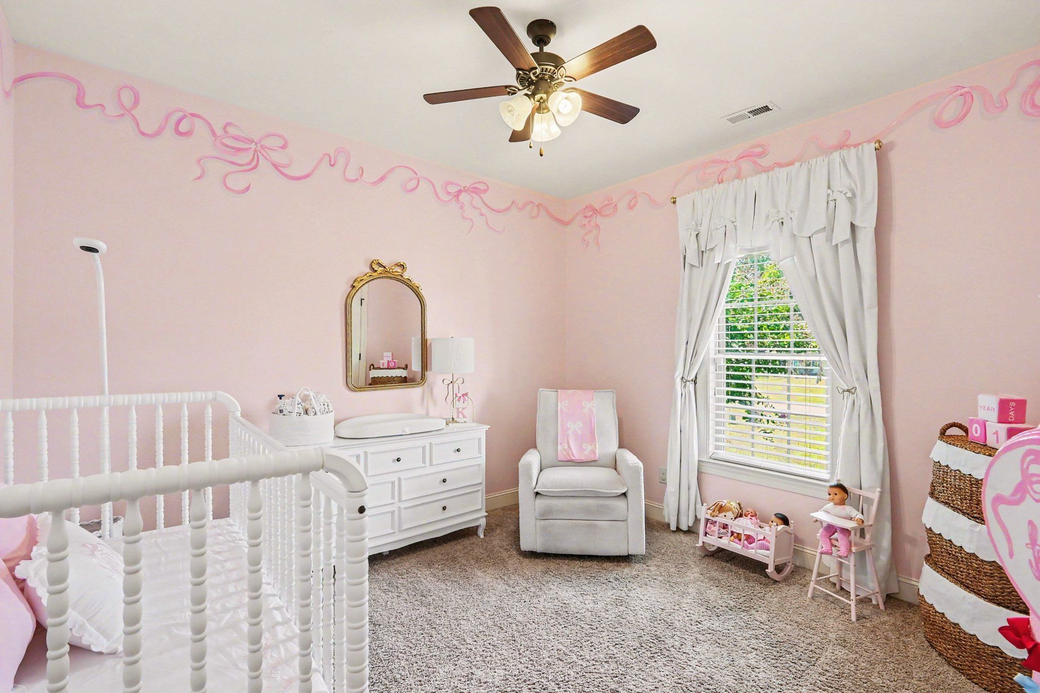 Pink nursery with a white crib, changing dresser, glider chair, and toys by a curtained window.