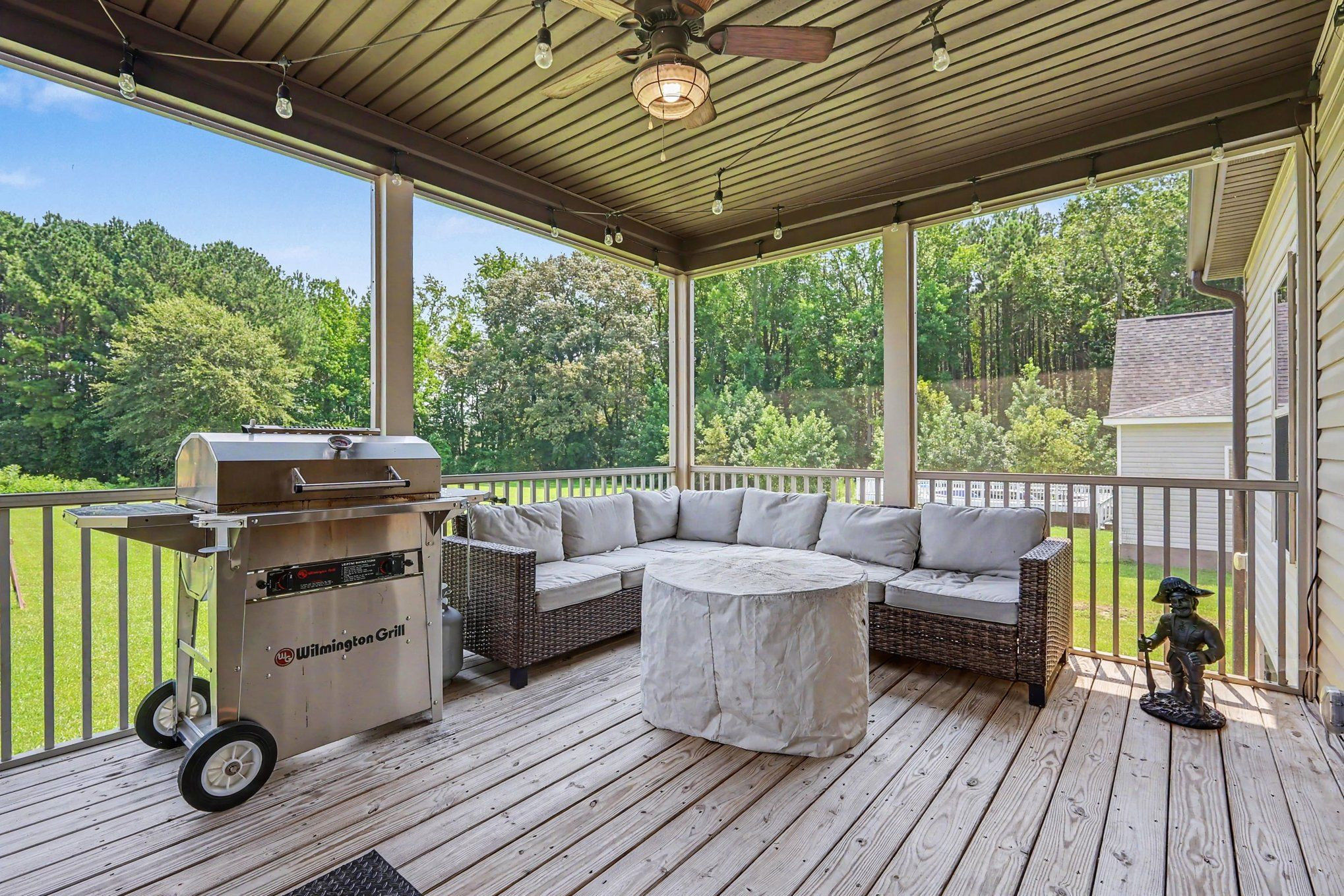 Covered deck with a stainless steel grill, wicker sectional, and wooded views.