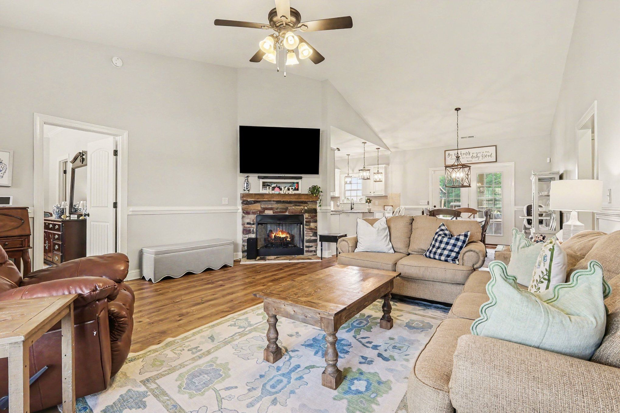 Furnished living room with a stone fireplace, wall-mounted television, and open kitchen and dining area.