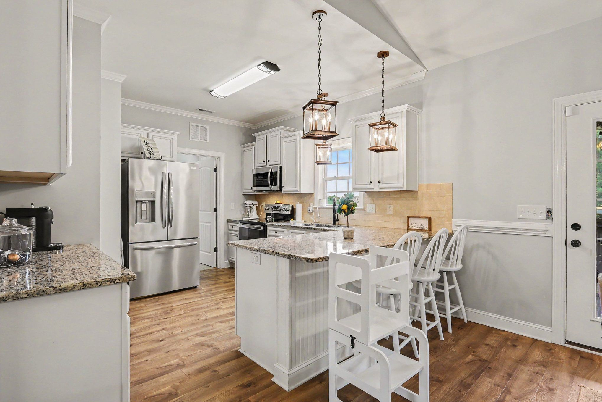 Kitchen with a granite island, white bar stools, stainless steel appliances, and warm wood flooring.