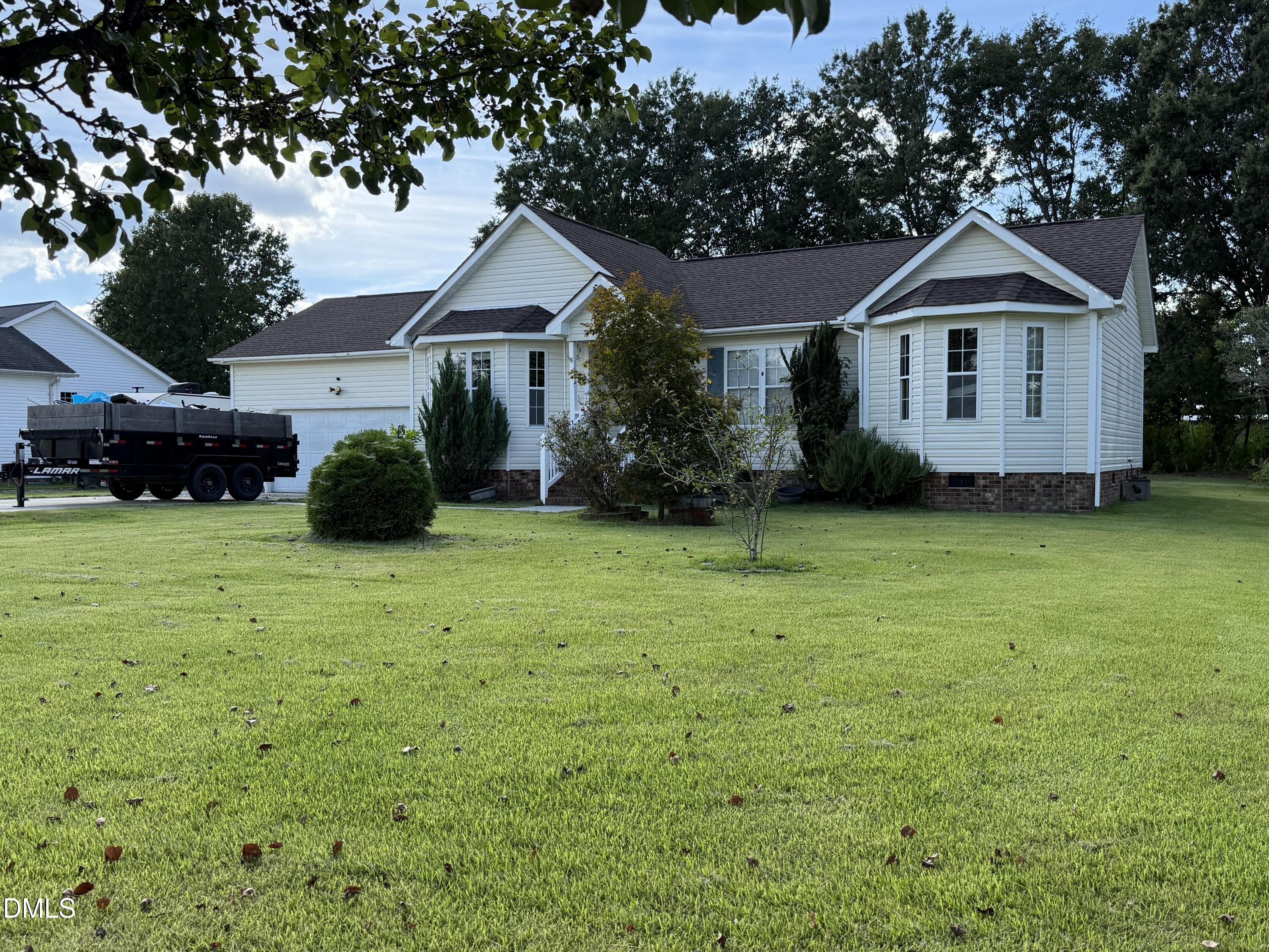 White single-story house with a brick foundation, broad lawn, and black dump trailer beside the garage.