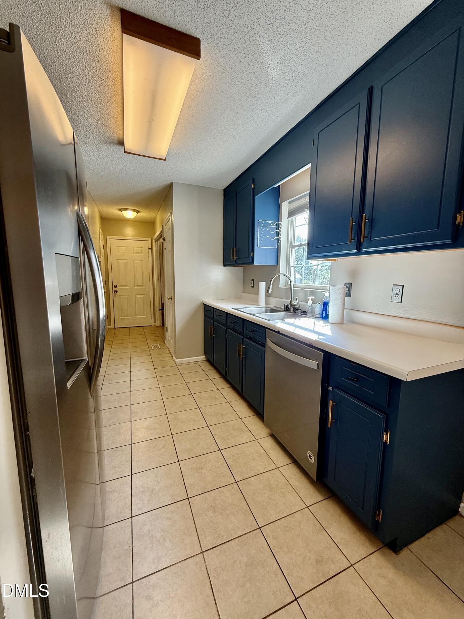 Galley kitchen with dark blue cabinets, stainless steel appliances, and a double-basin sink beneath a window.