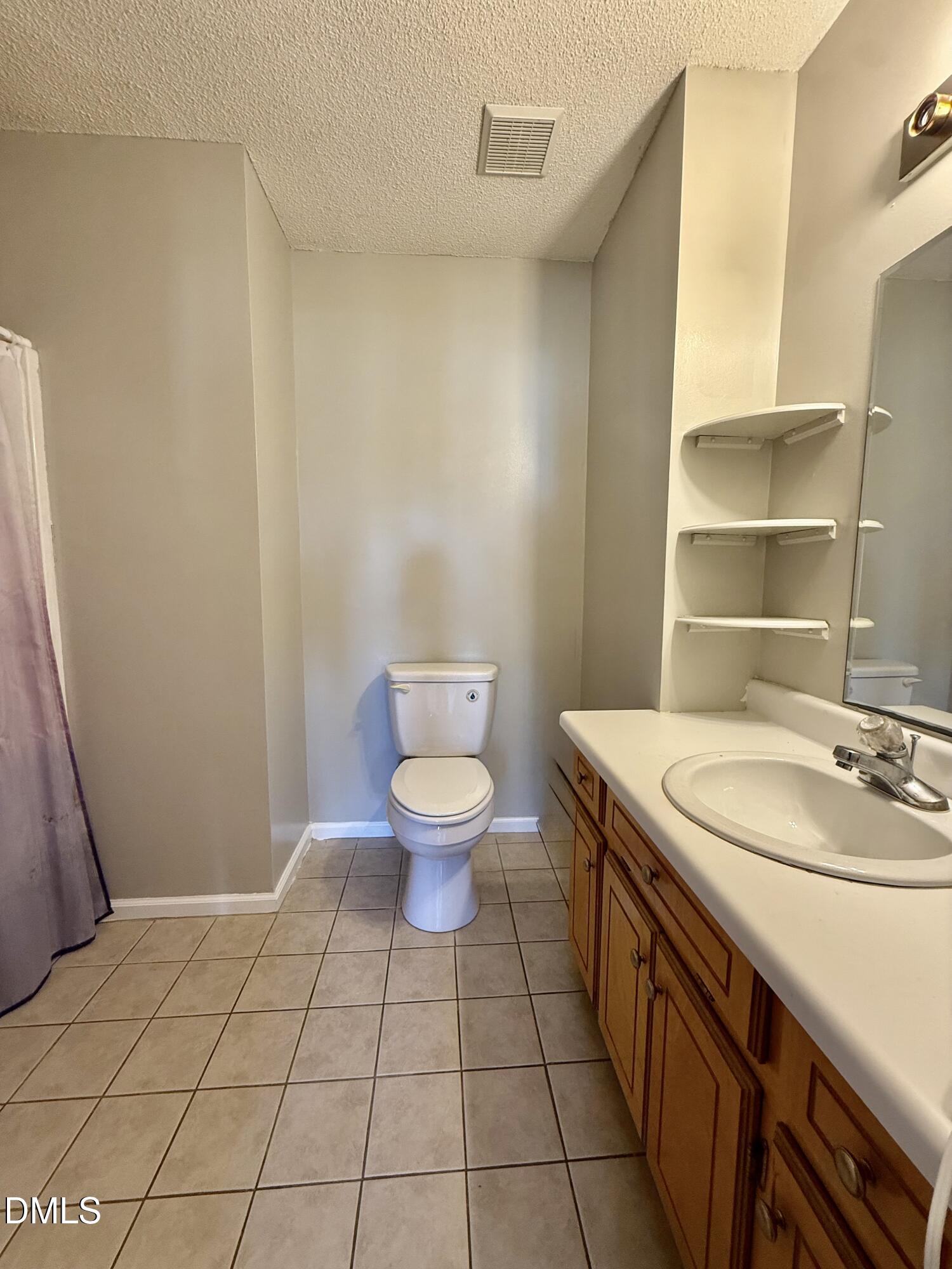 Bathroom with a tiled floor, white toilet, wood vanity, and corner shelves beside a large mirror.