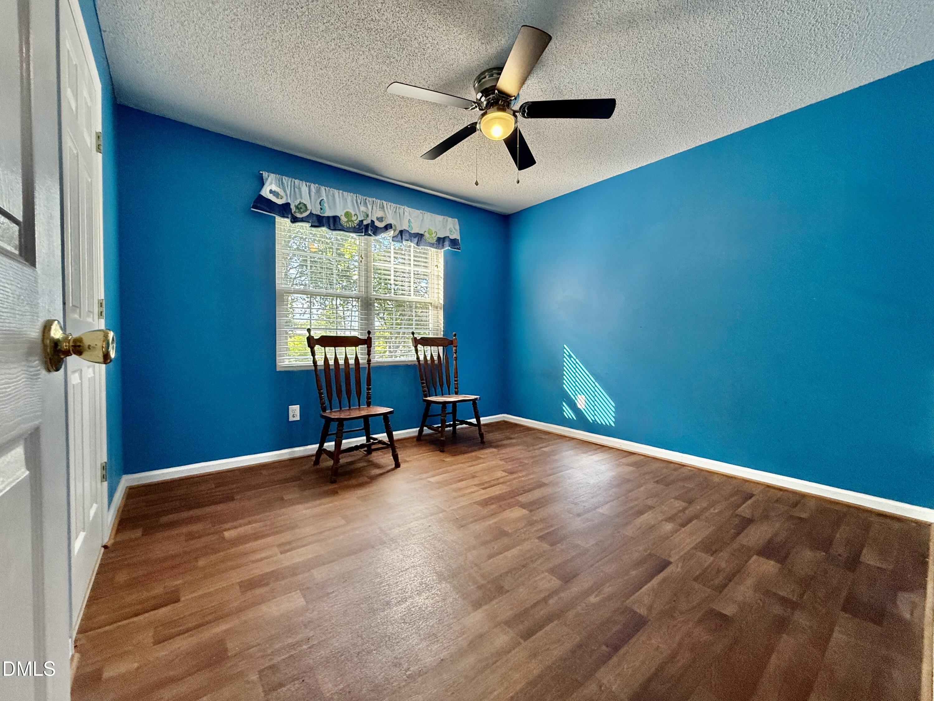 Bright blue room with wood flooring, two chairs beneath a window, and a ceiling fan.