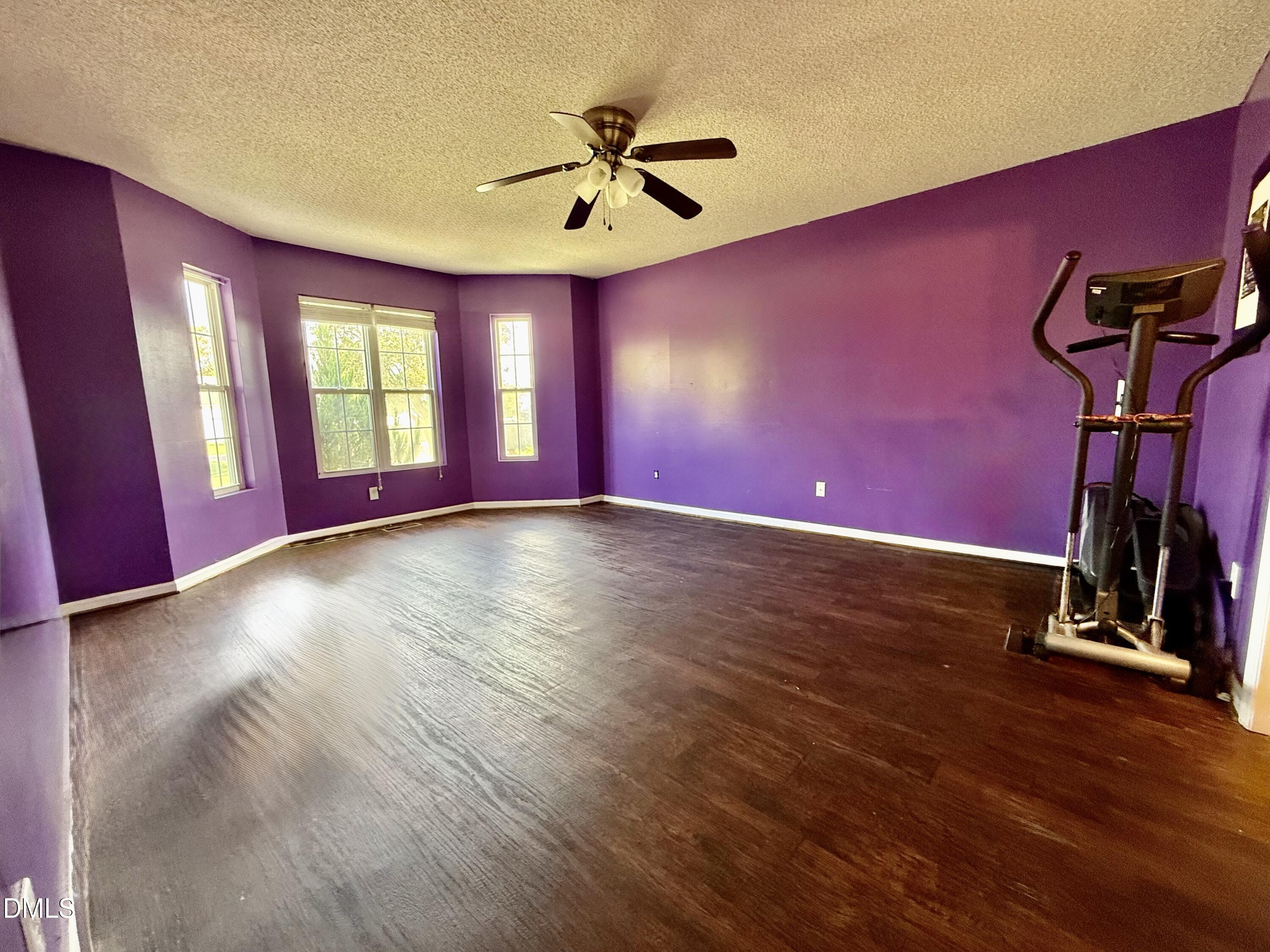 Mostly empty purple room with dark wood floors, three windows, and an elliptical machine.