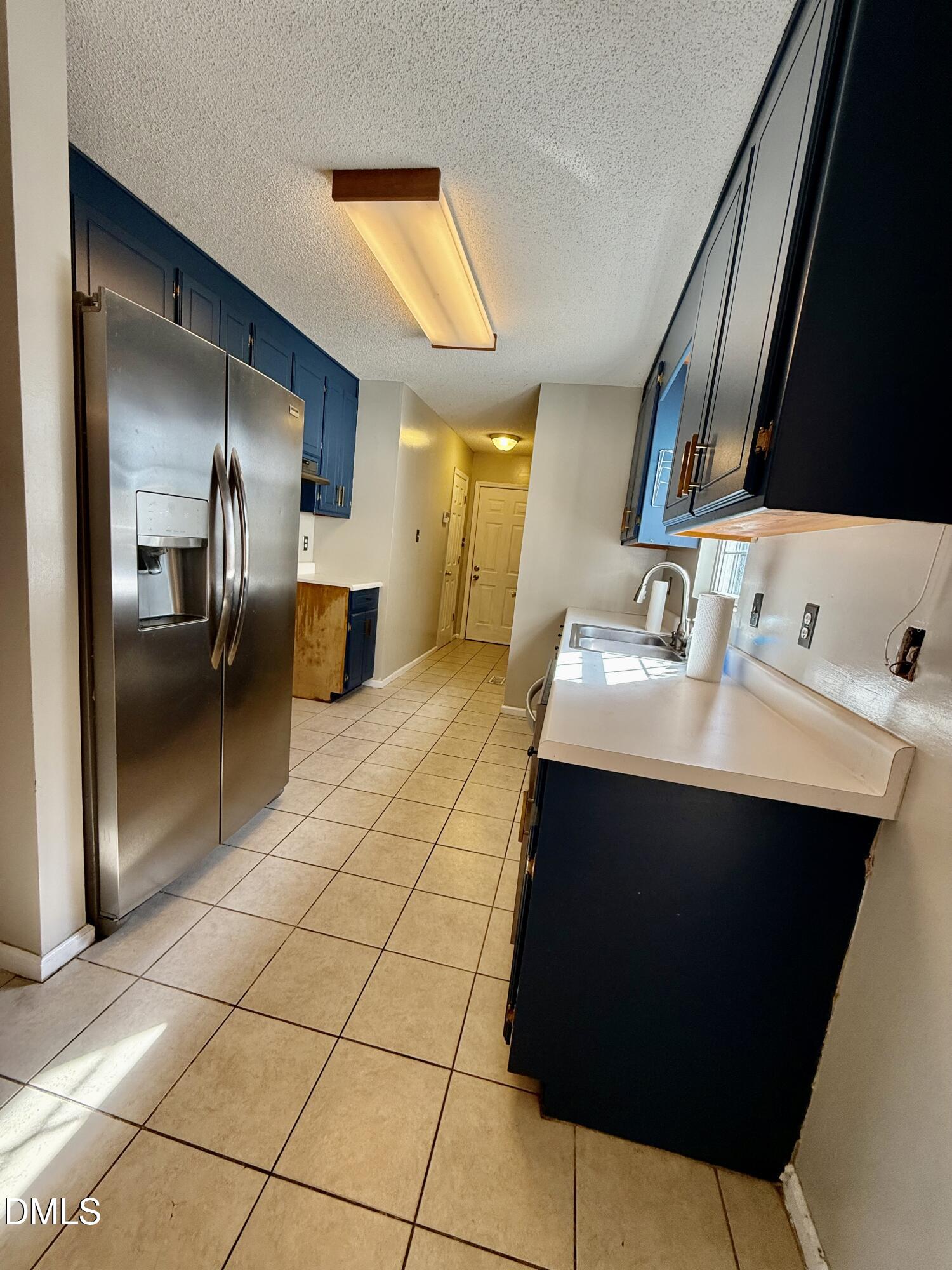 Galley kitchen with dark blue cabinets, stainless steel refrigerator, and beige tile floor.