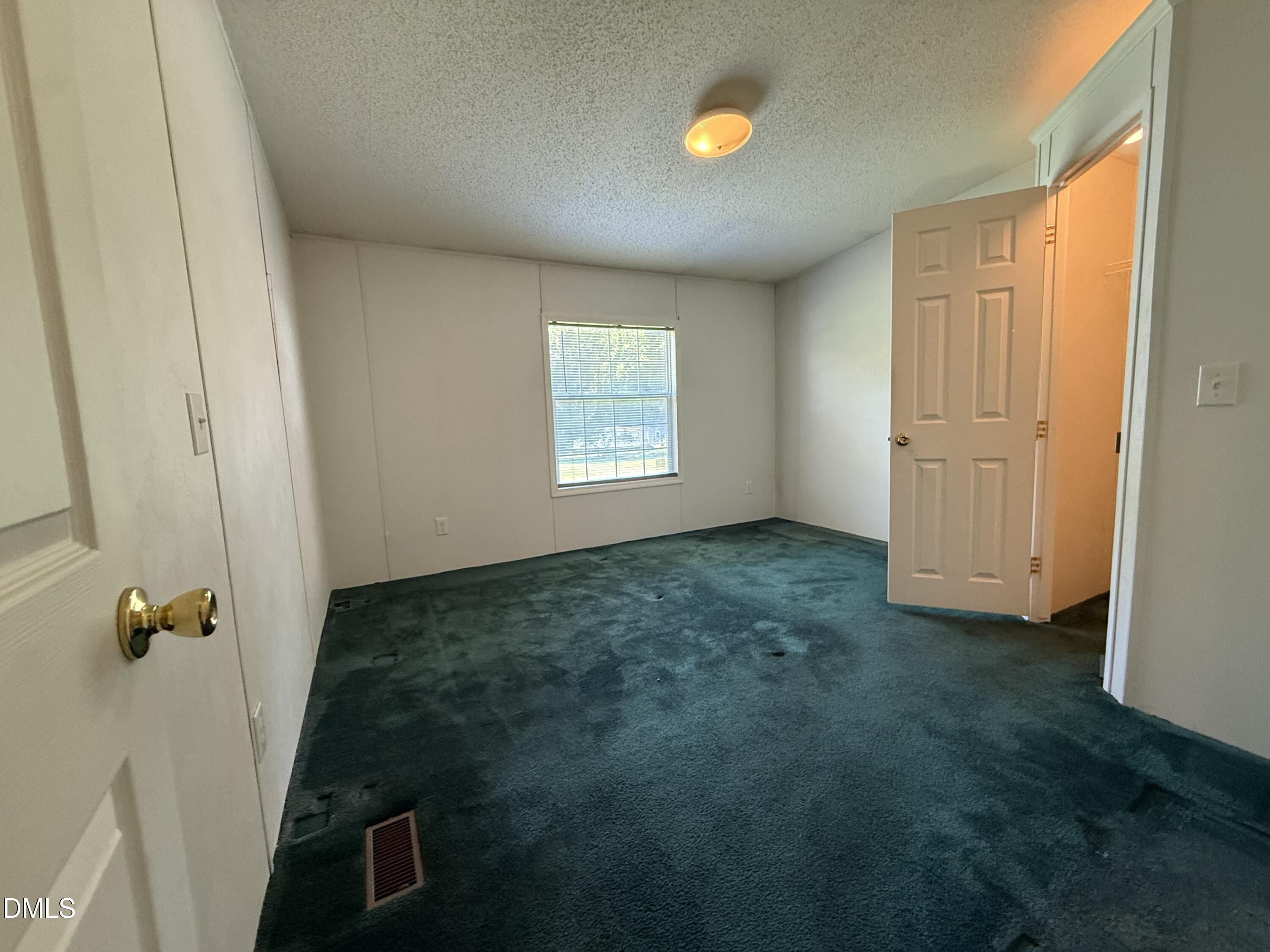 Empty bedroom with dark teal carpet, a window with blinds, and an open door.
