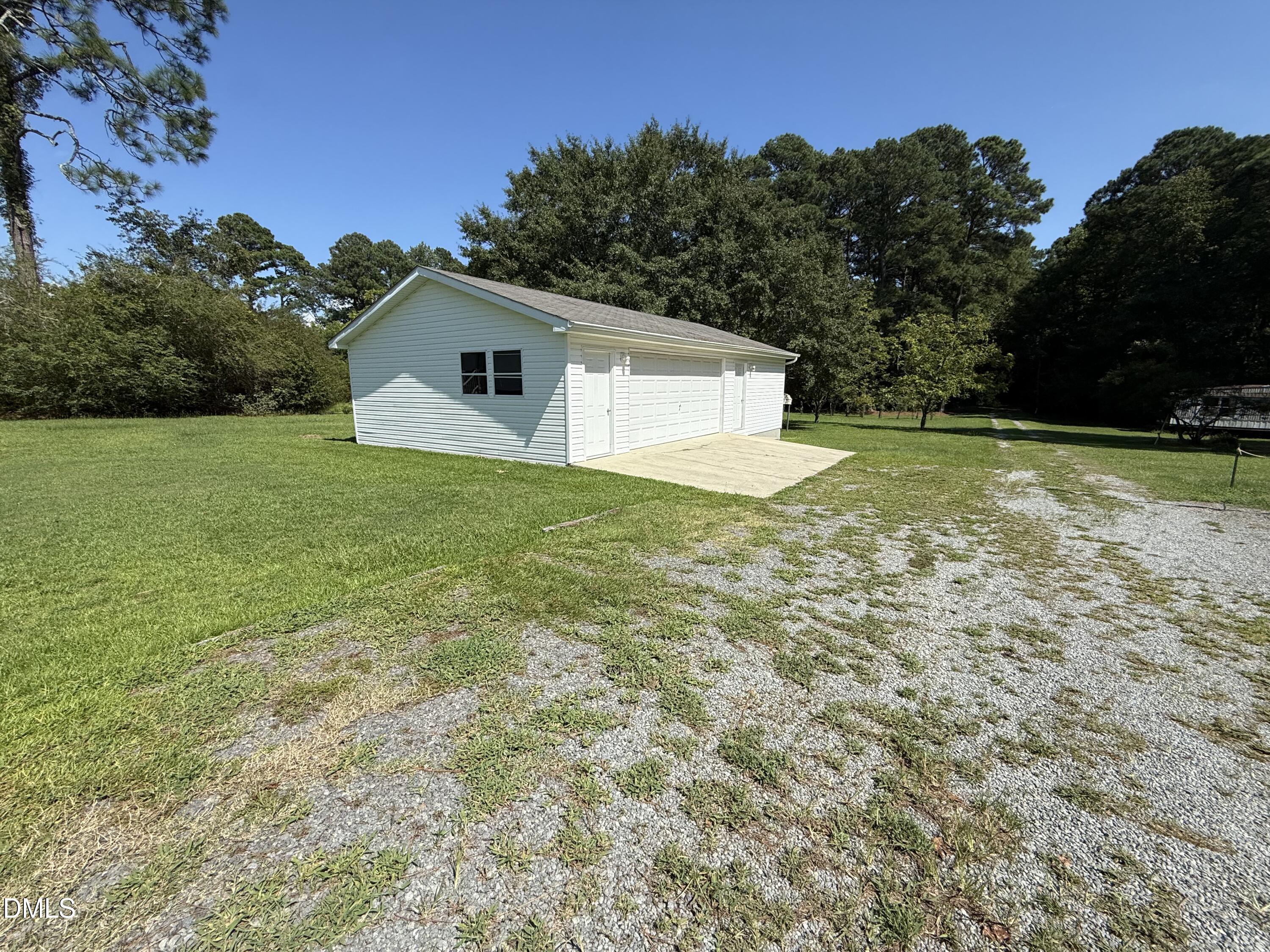 White detached garage with multiple doors and a concrete apron beside a gravel drive on a wooded lot.