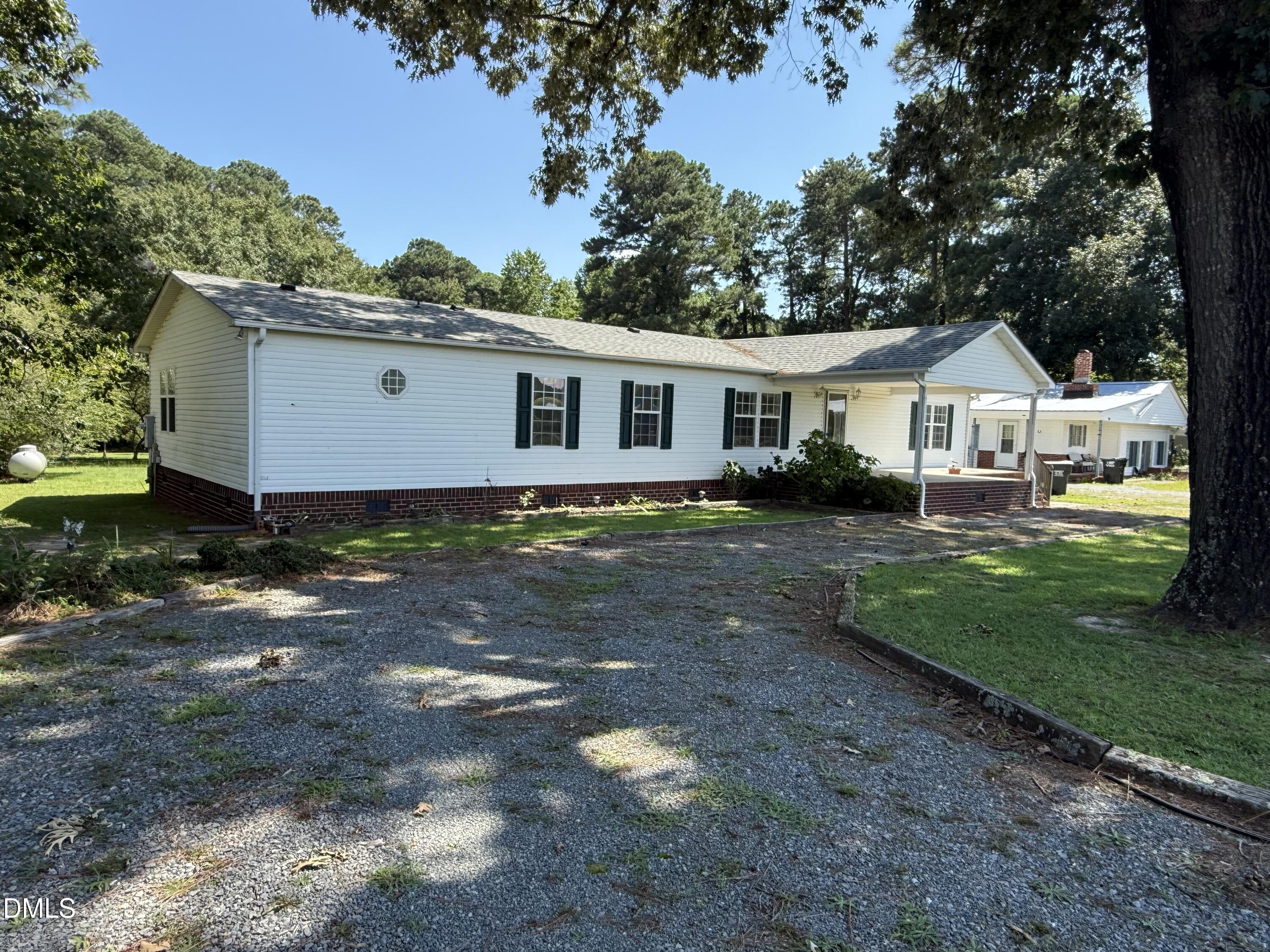 White single-story house with dark shutters, brick foundation, and covered porch beside a gravel driveway.