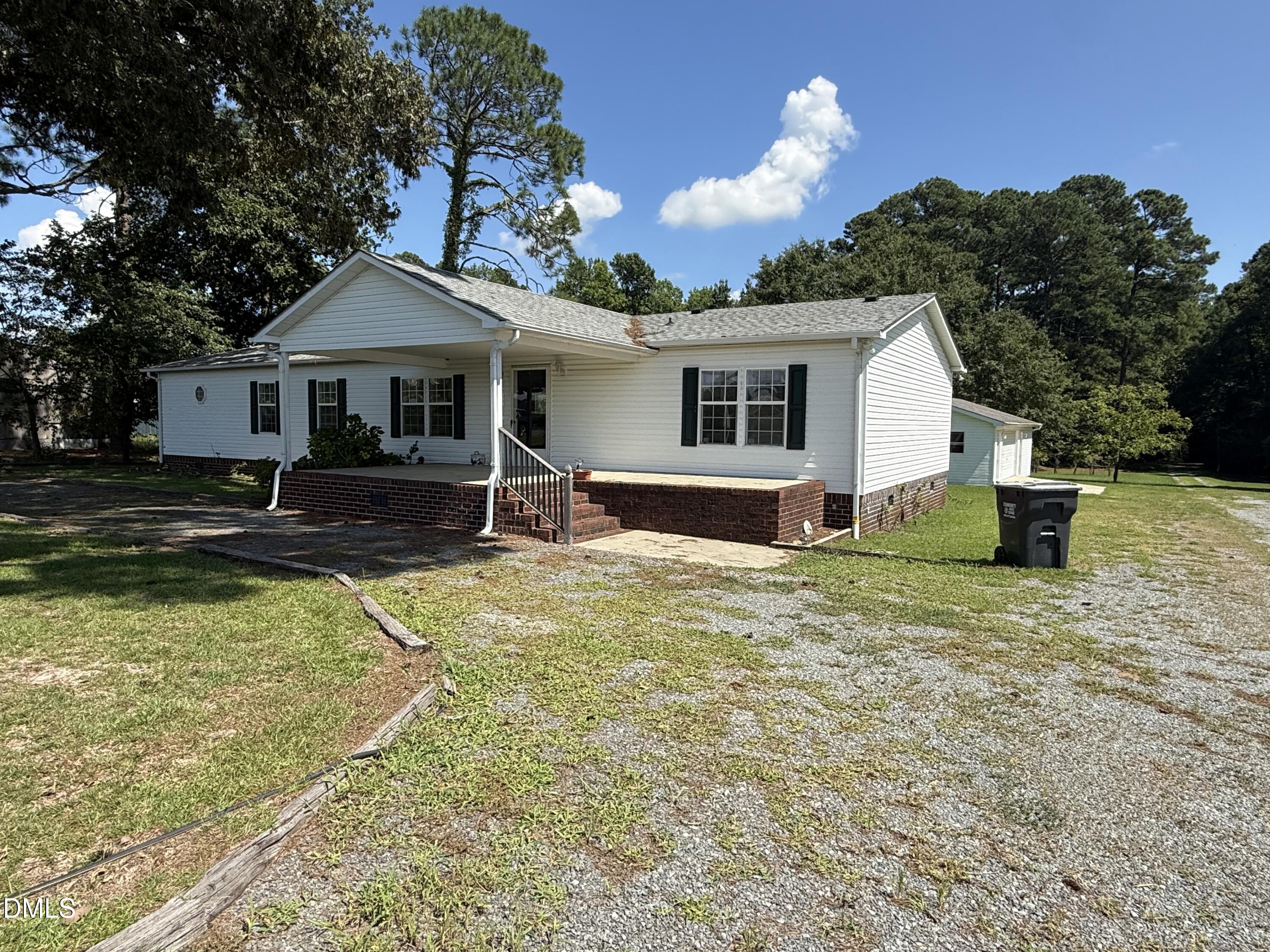 White single-story house with a raised brick porch, gravel driveway, and mature trees.