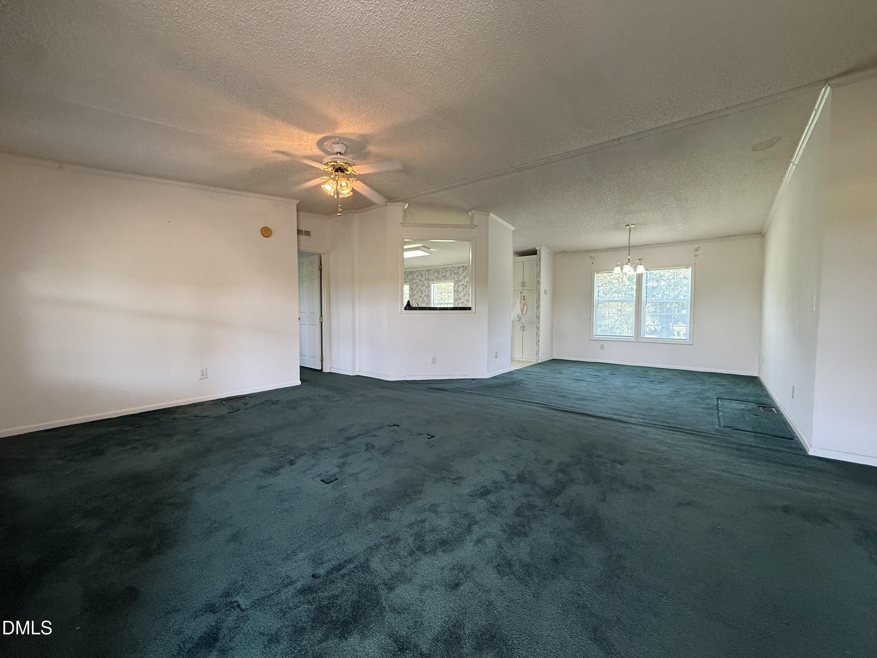 Empty living and dining area with dark teal carpet, ceiling fan, and large window.