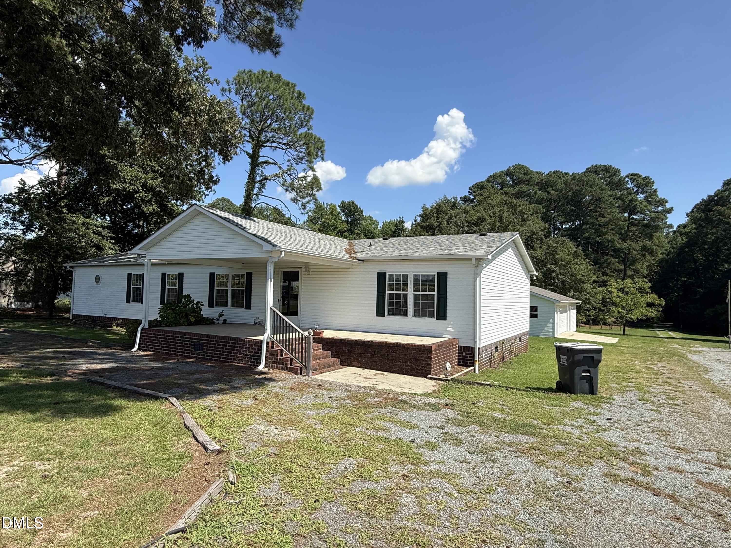 White single-story home with a brick porch, gravel driveway, and wooded surroundings.