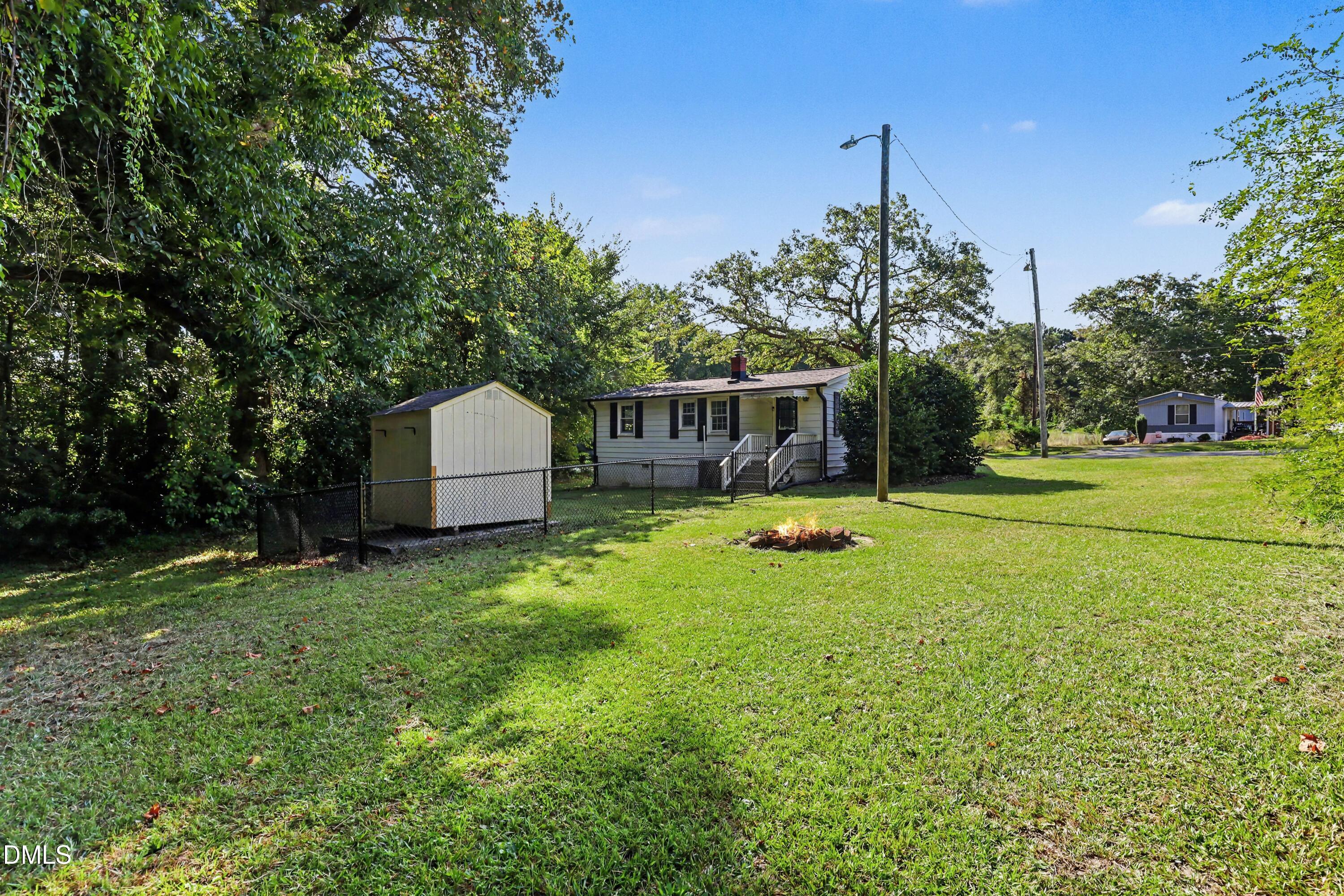Single-story house beyond a grassy yard, with a shed, chain-link fence, and fire pit.
