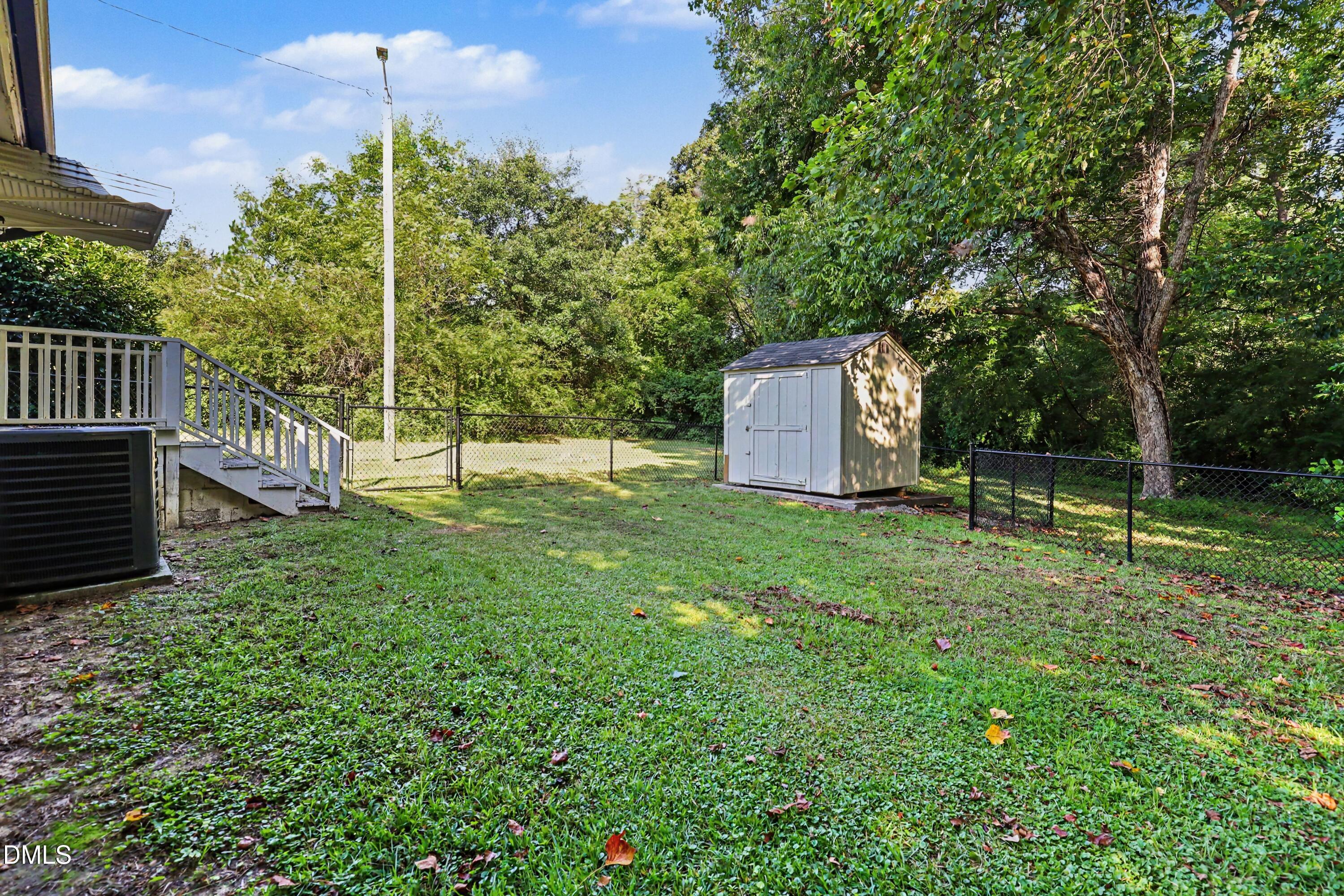 Fenced grassy backyard with a white storage shed, mature trees, and stairs from the rear deck.