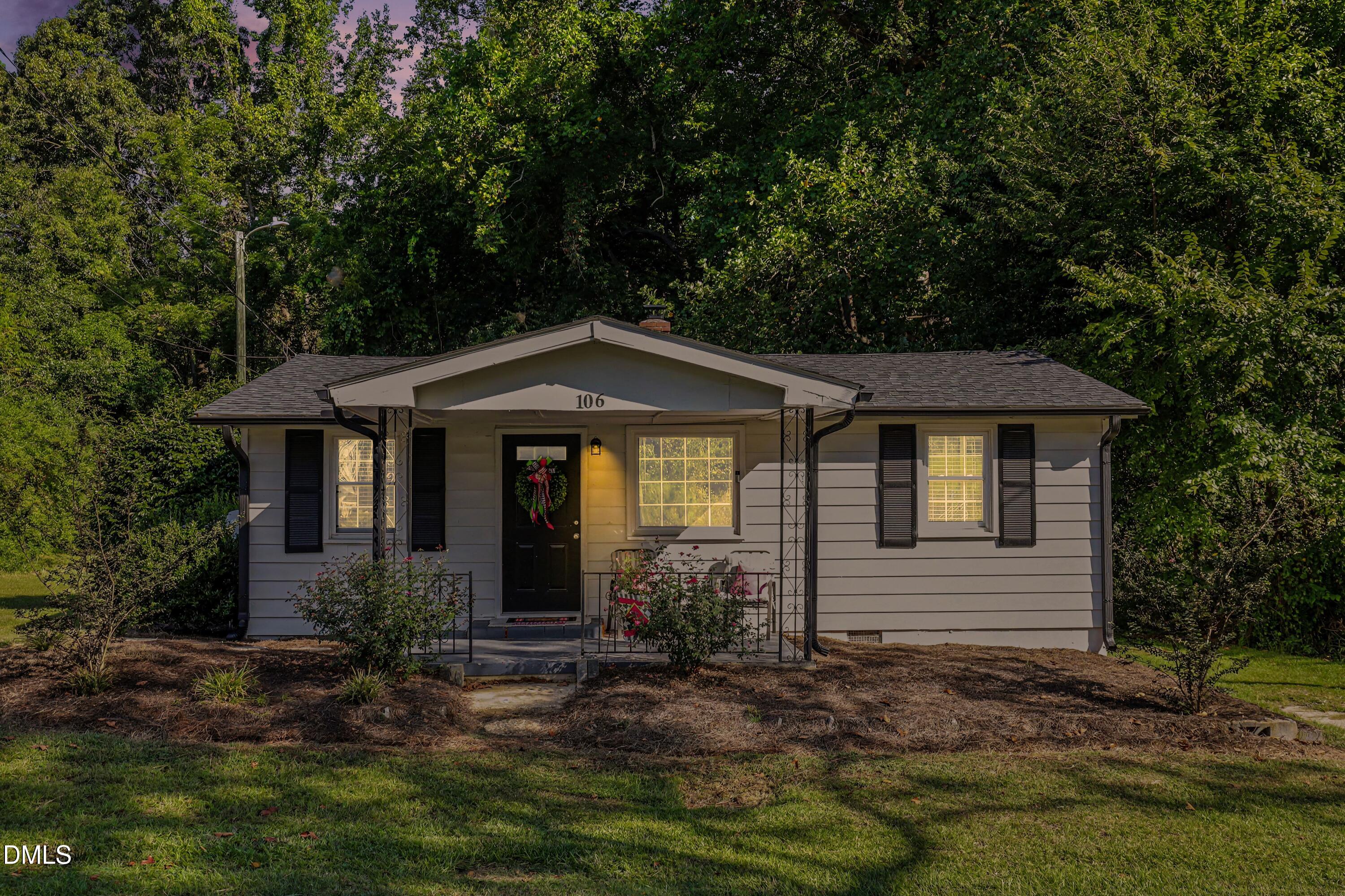 Gray single-story home with black shutters, a covered porch, and mature trees behind it.
