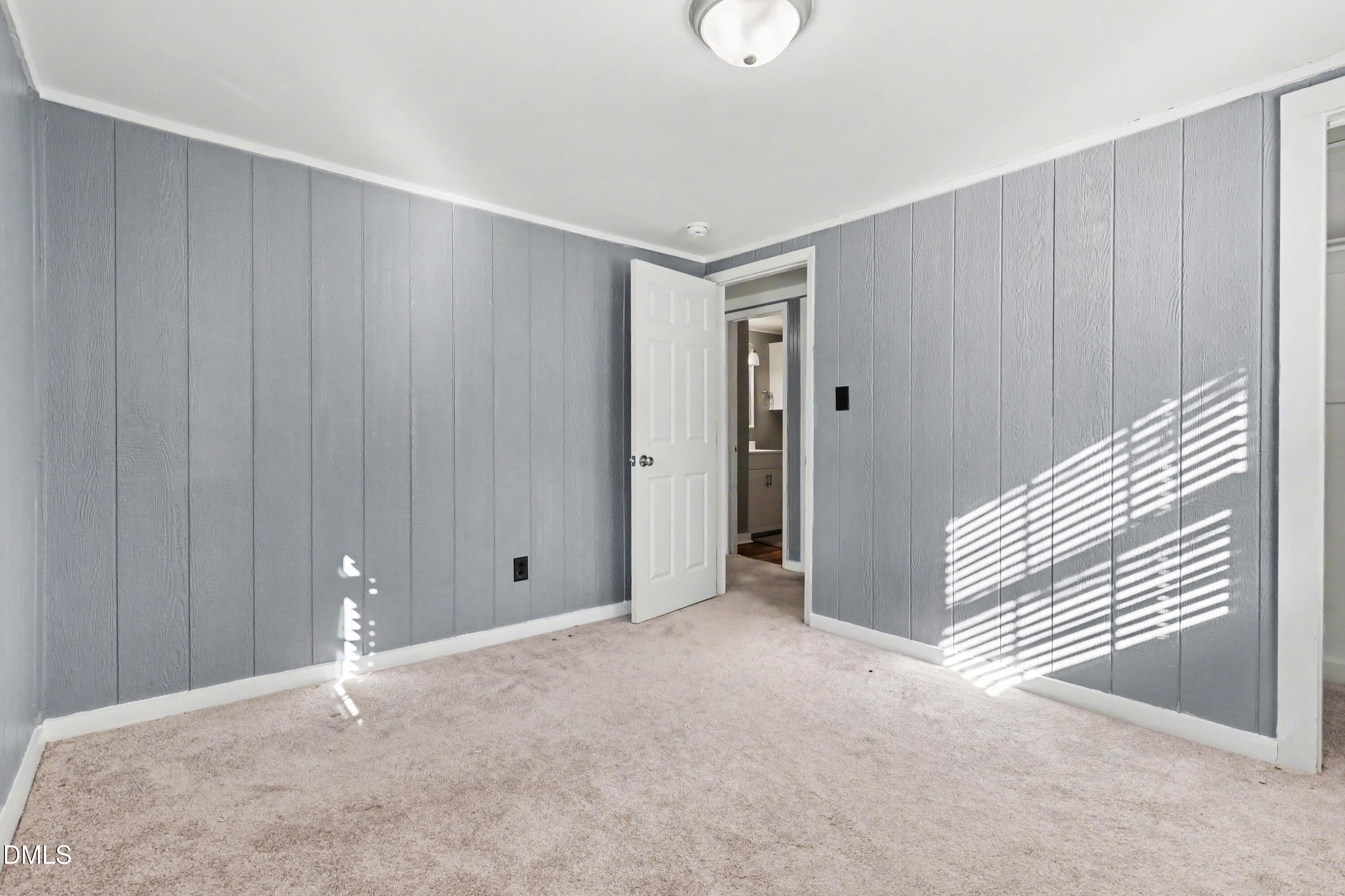 Vacant bedroom with gray paneled walls, beige carpet, and sunlight casting striped shadows.