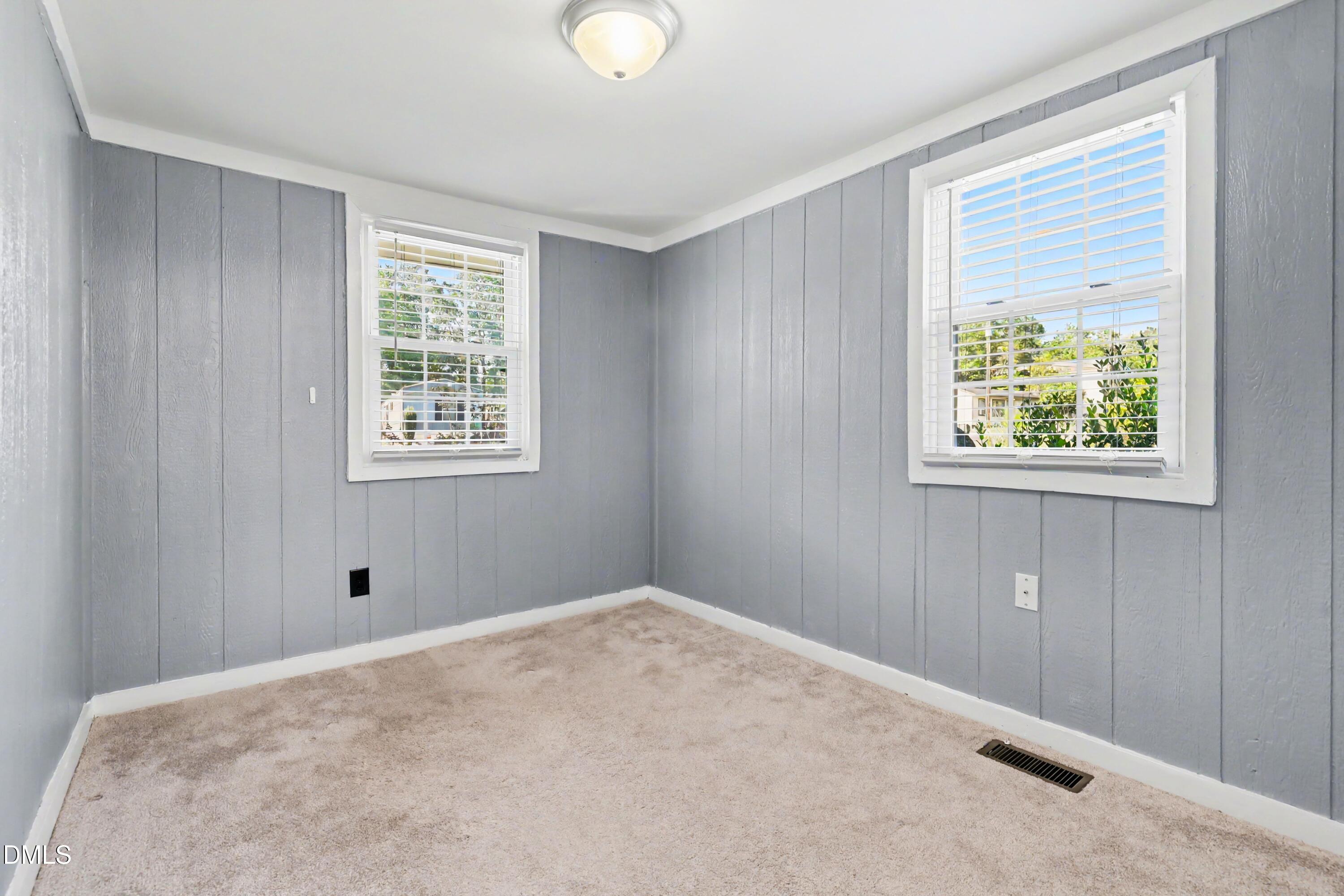 Empty room with gray paneled walls, beige carpet, and two windows with white blinds.