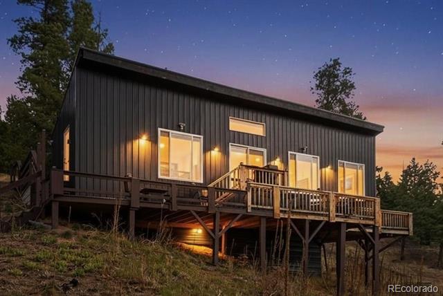 Modern dark-sided home with a raised wooden deck and glowing windows at dusk.