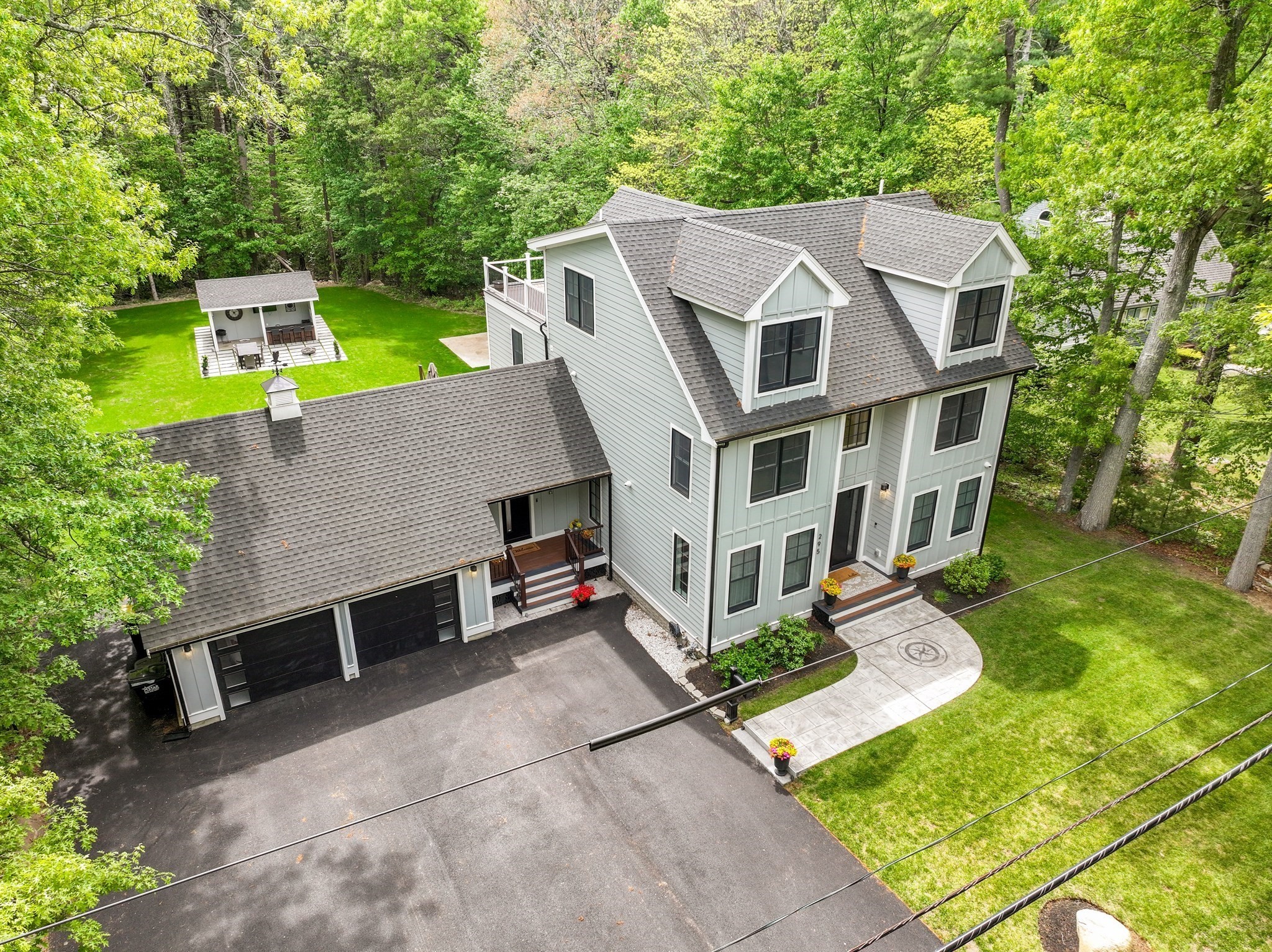 Aerial view of a light-gray home with a large garage, backyard pavilion, and wooded lawn.