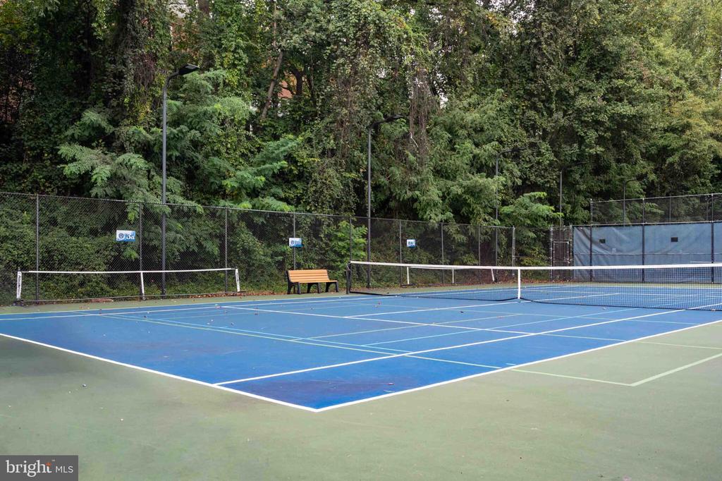 Outdoor tennis courts with blue playing surfaces, nets, chain-link fencing, and a bench.