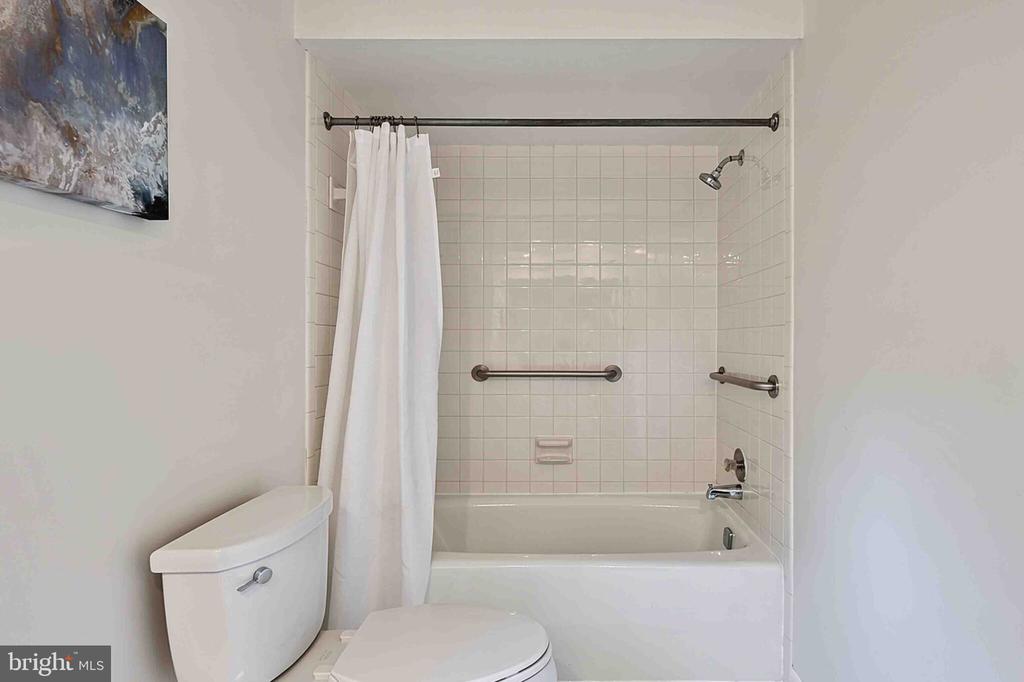 Bathroom with a tiled tub and shower, white curtain, grab bars, and toilet.