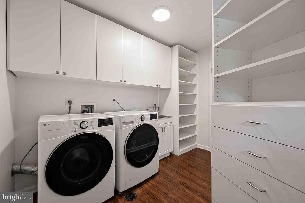 Laundry room with front-loading washer and dryer, white cabinetry, open shelves, and wood-look flooring.