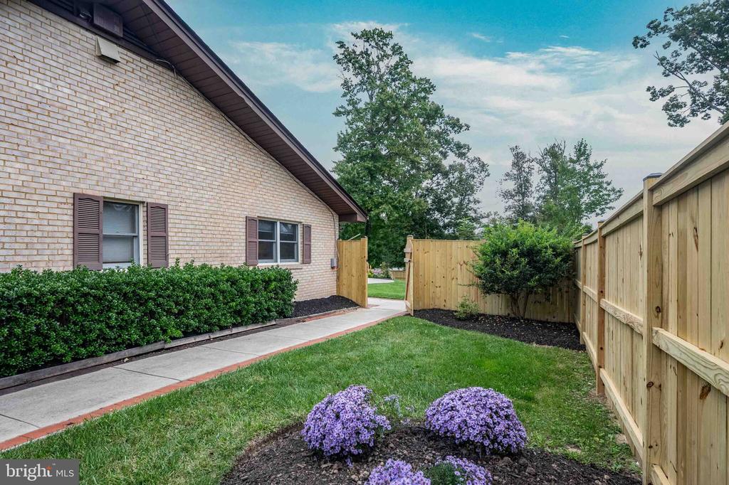 Brick home’s side yard with a concrete walkway, wooden fencing, and purple flowers.
