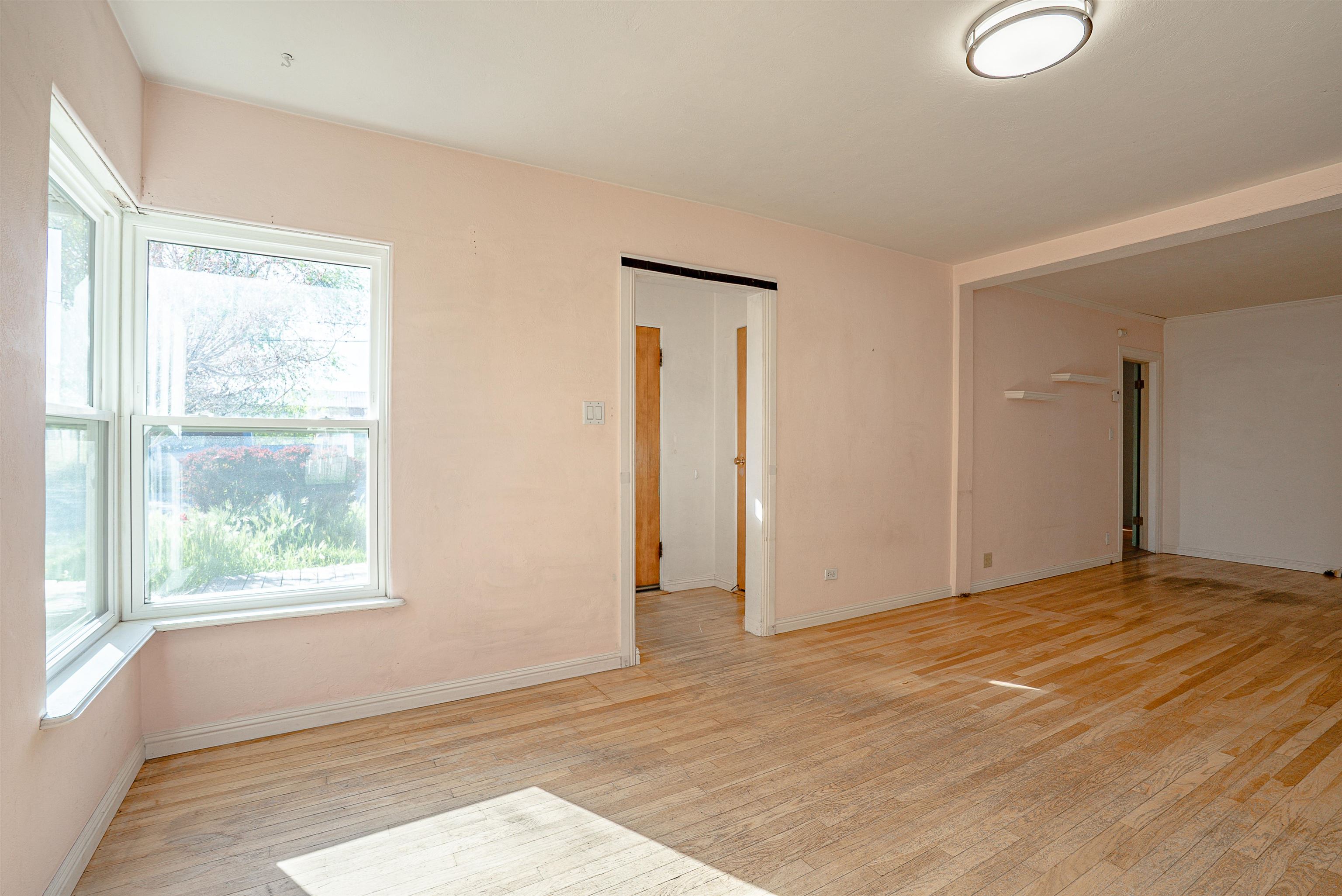Vacant room with hardwood floors, pale pink walls, a large corner window, and open doorways.